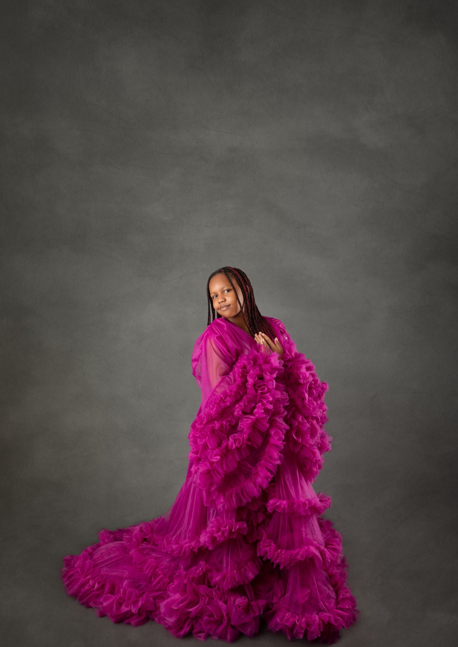 Young girl in dramatic magenta ruffle tulle gown poses elegantly against gray studio backdrop.