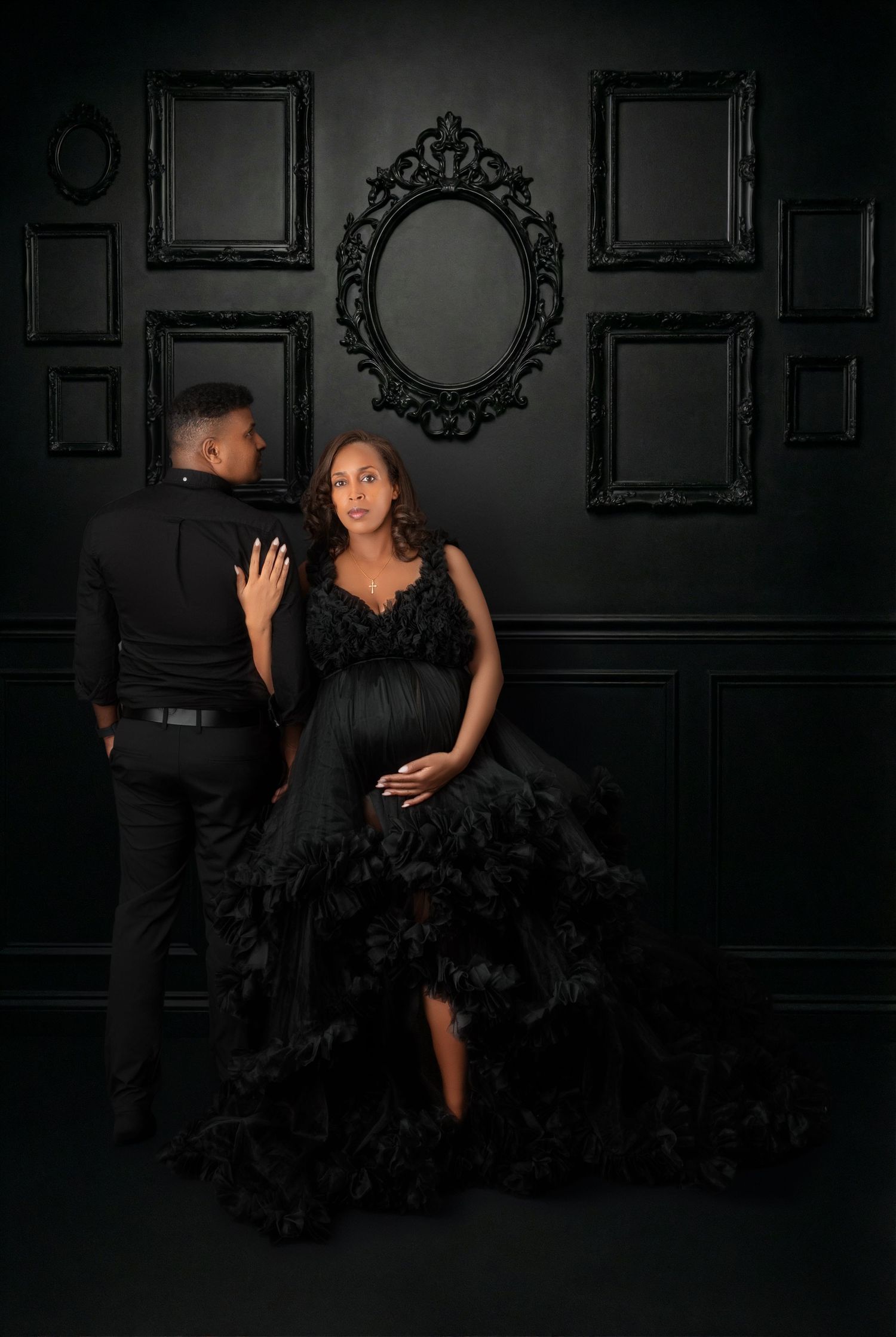 Couple in matching black outfits pose against dramatic black wall with ornate frames.