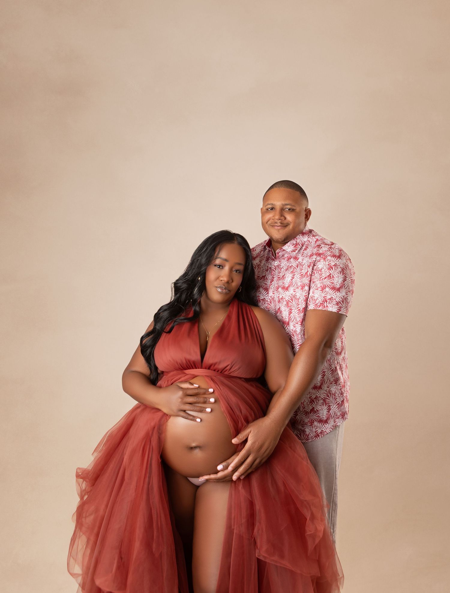 Couple tenderly cradles pregnant belly together in matching red and floral outfits in studio.