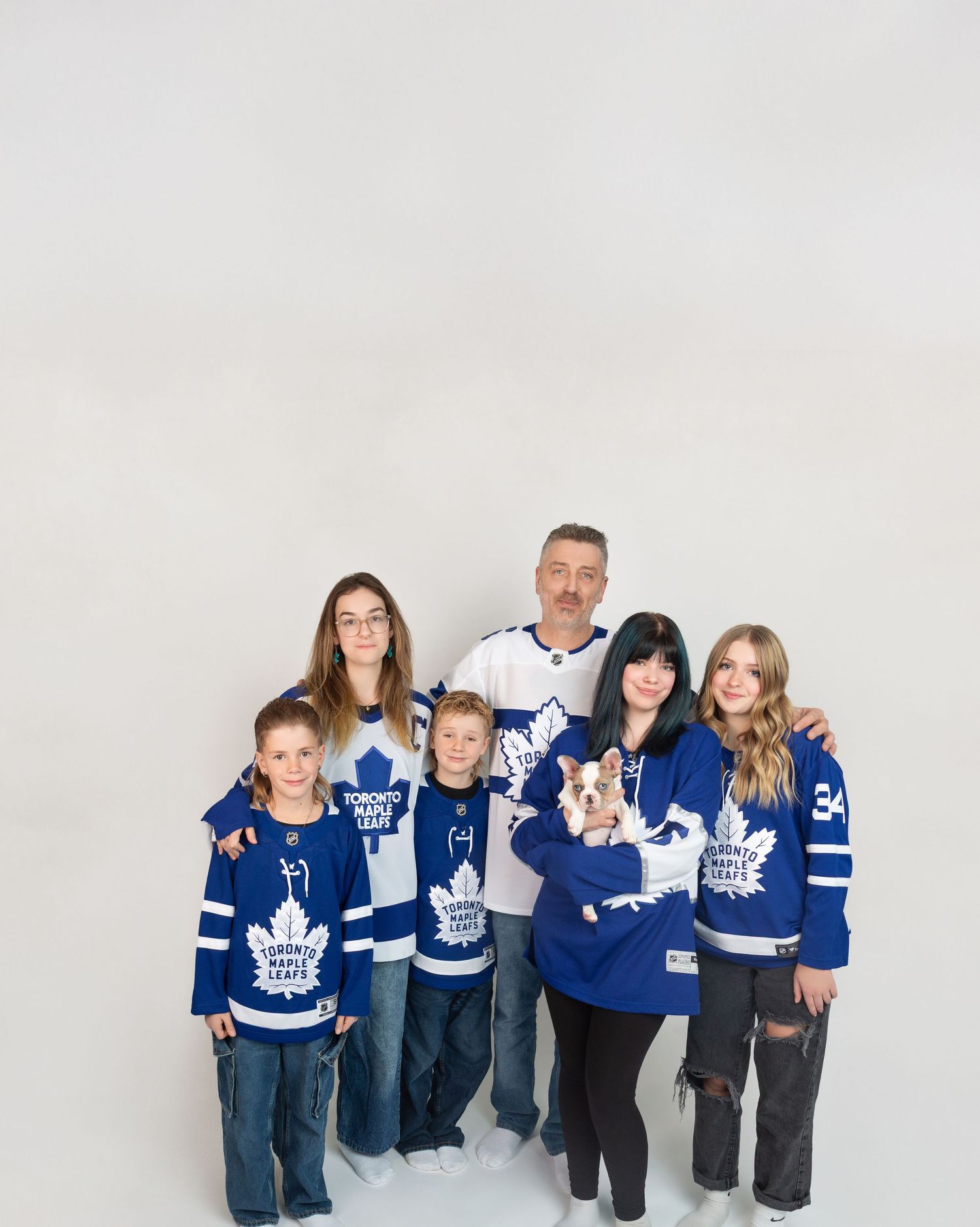 Family of six wearing Toronto Maple Leafs jerseys poses together holding a small dog in a studio.