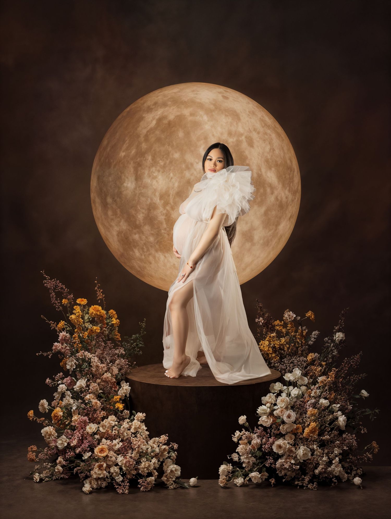 Pregnant woman in white gown poses on pedestal with moon backdrop and floral arrangements in elegant maternity portrait.