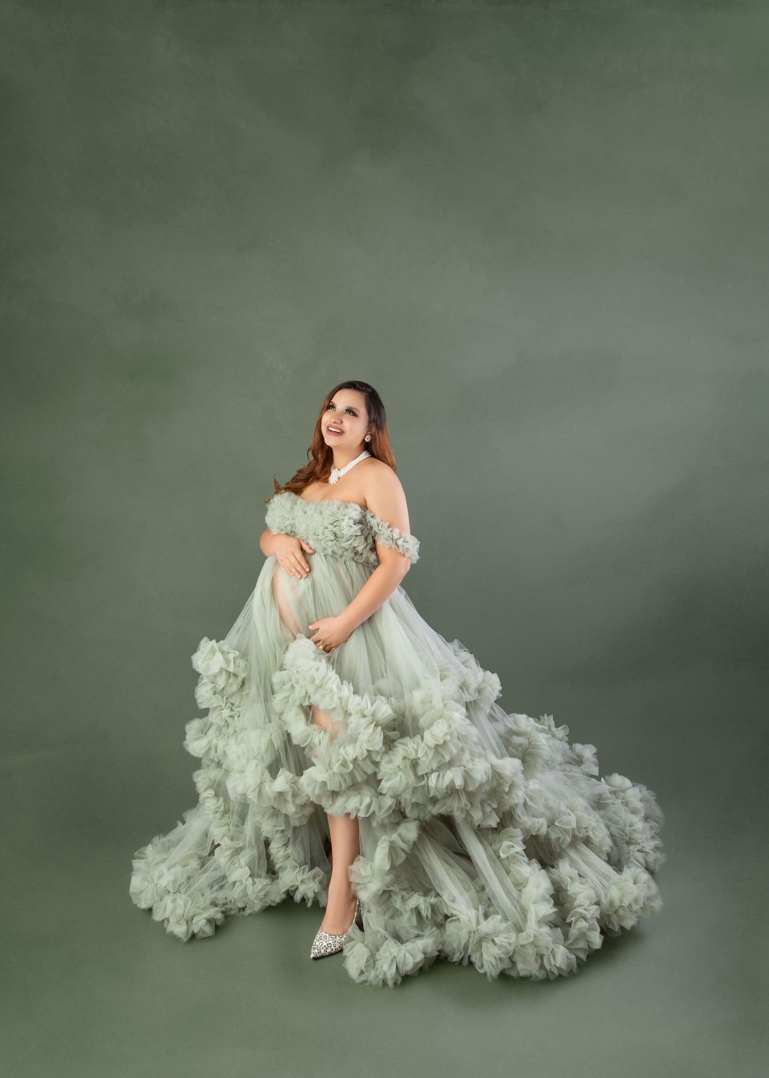 Pregnant woman in elegant sage green ruffled tulle maternity gown posing gracefully in studio.