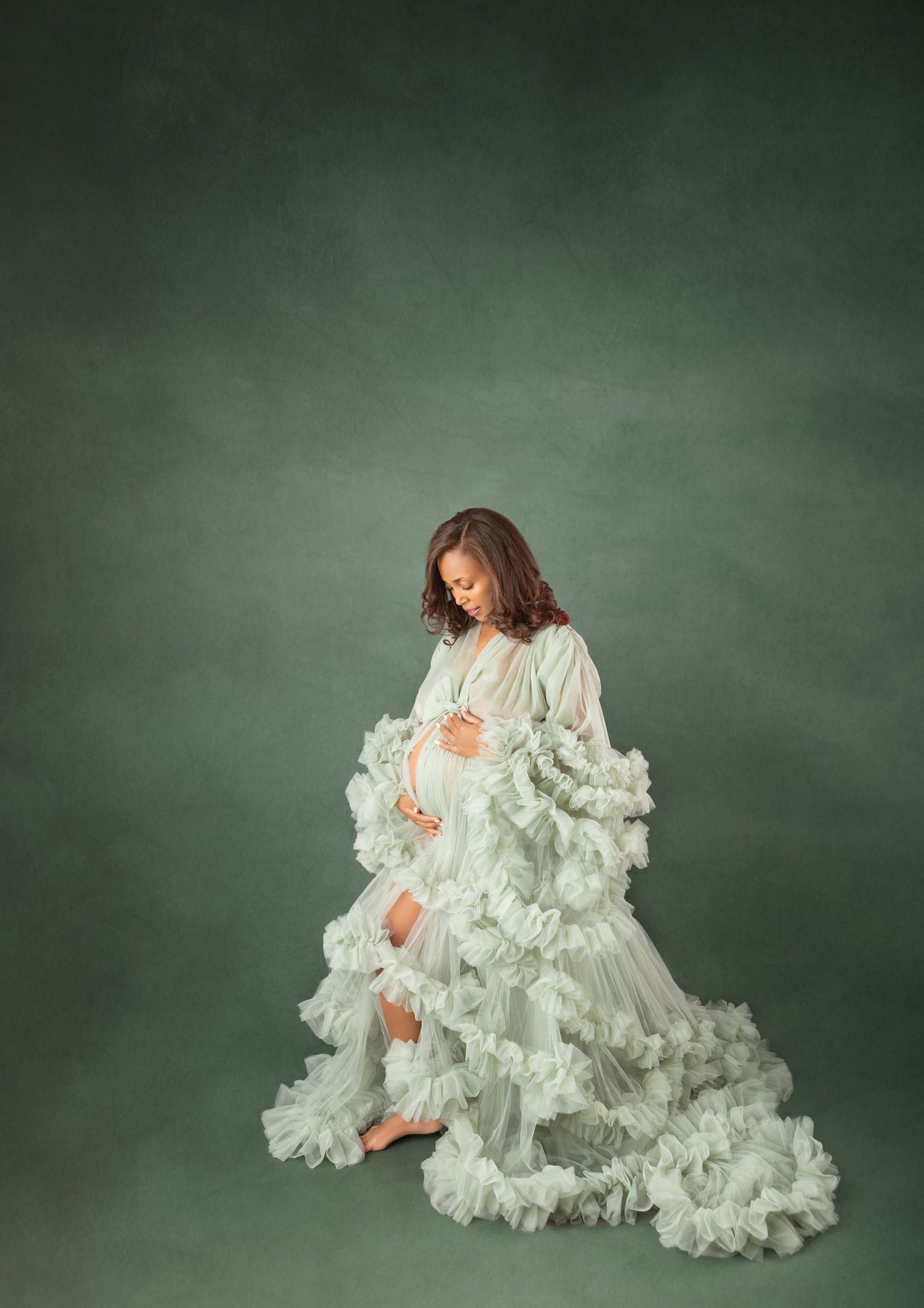 Pregnant woman in mint green ruffled tulle maternity gown cradles baby bump in moody green studio setting.