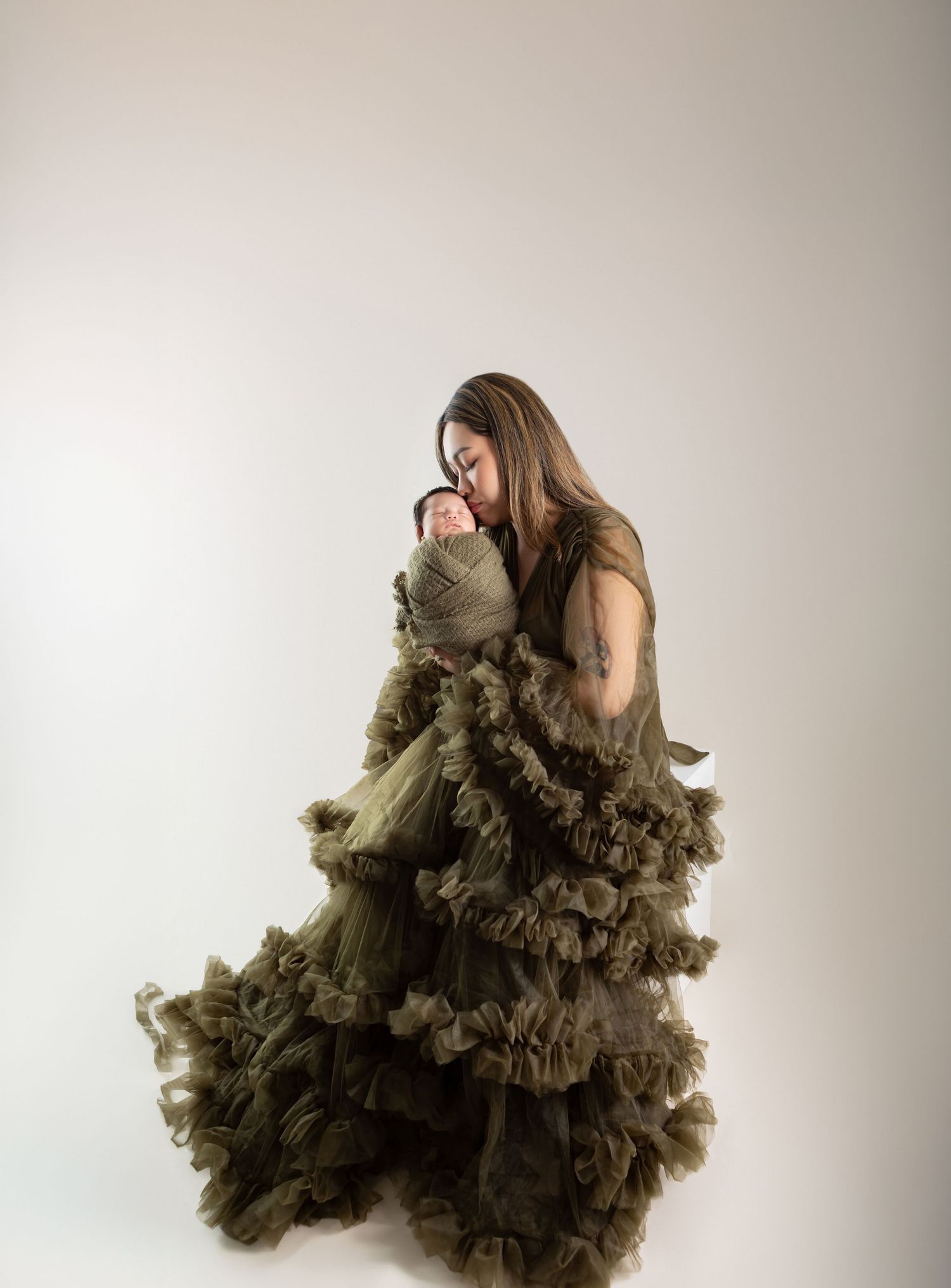 Woman in flowing olive ruffled maternity gown tenderly holds swaddled newborn in studio portrait.