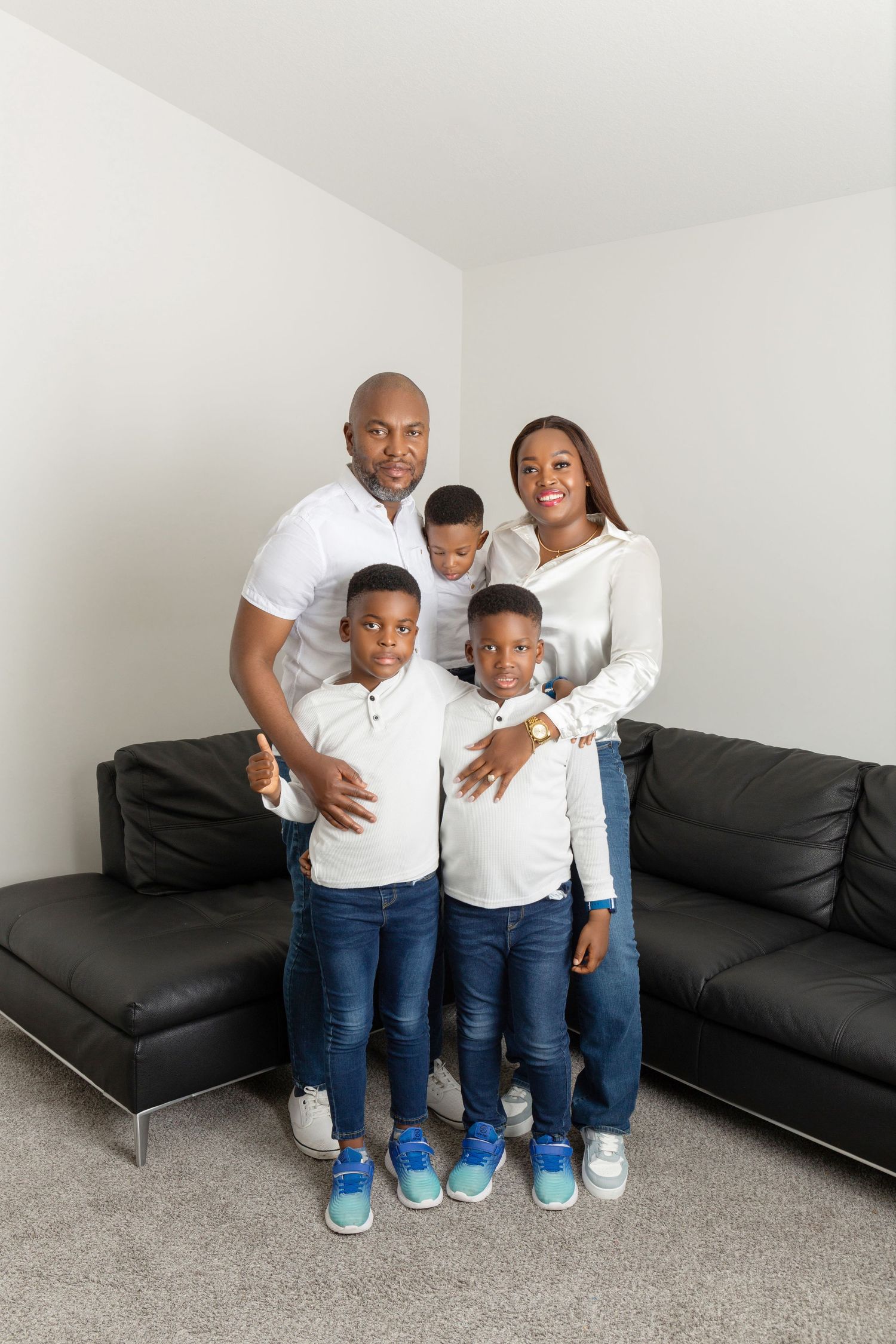 Happy Black family of five posing together in living room, parents with three young sons wearing matching white shirts and jeans.