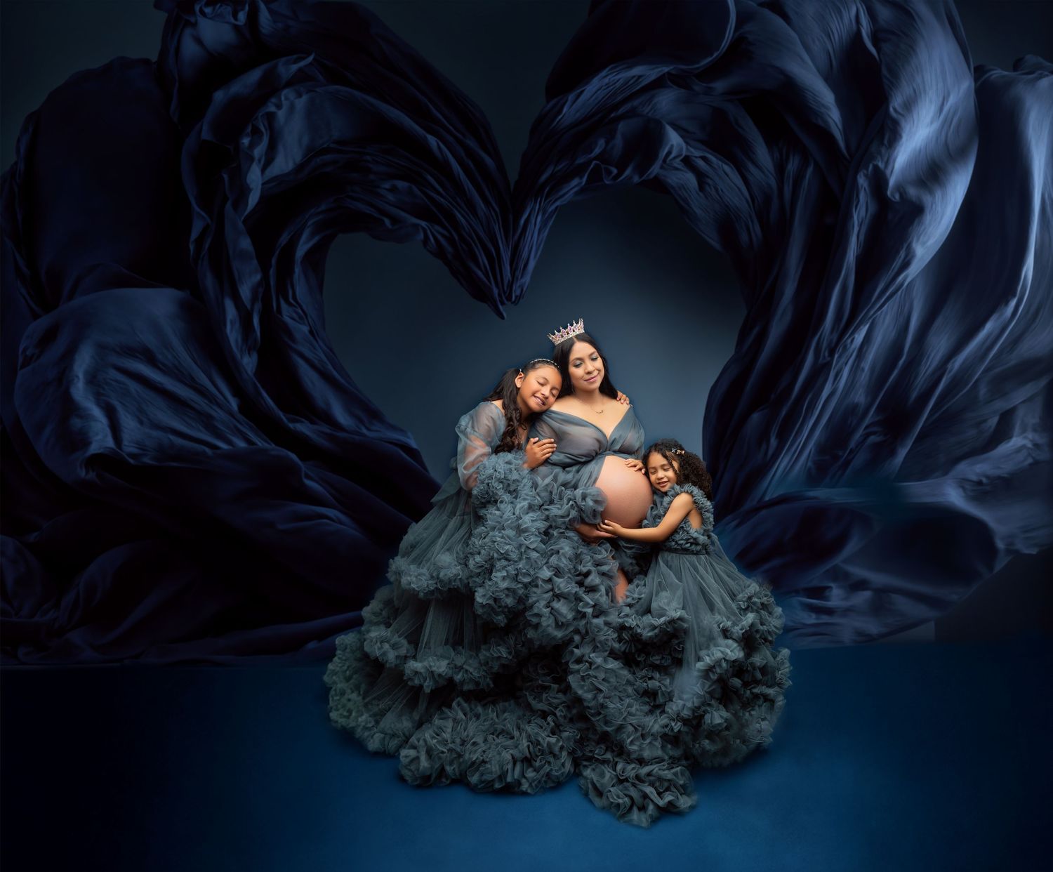 Pregnant woman in teal ruffle gown poses with children against dark blue flowing fabric heart backdrop.