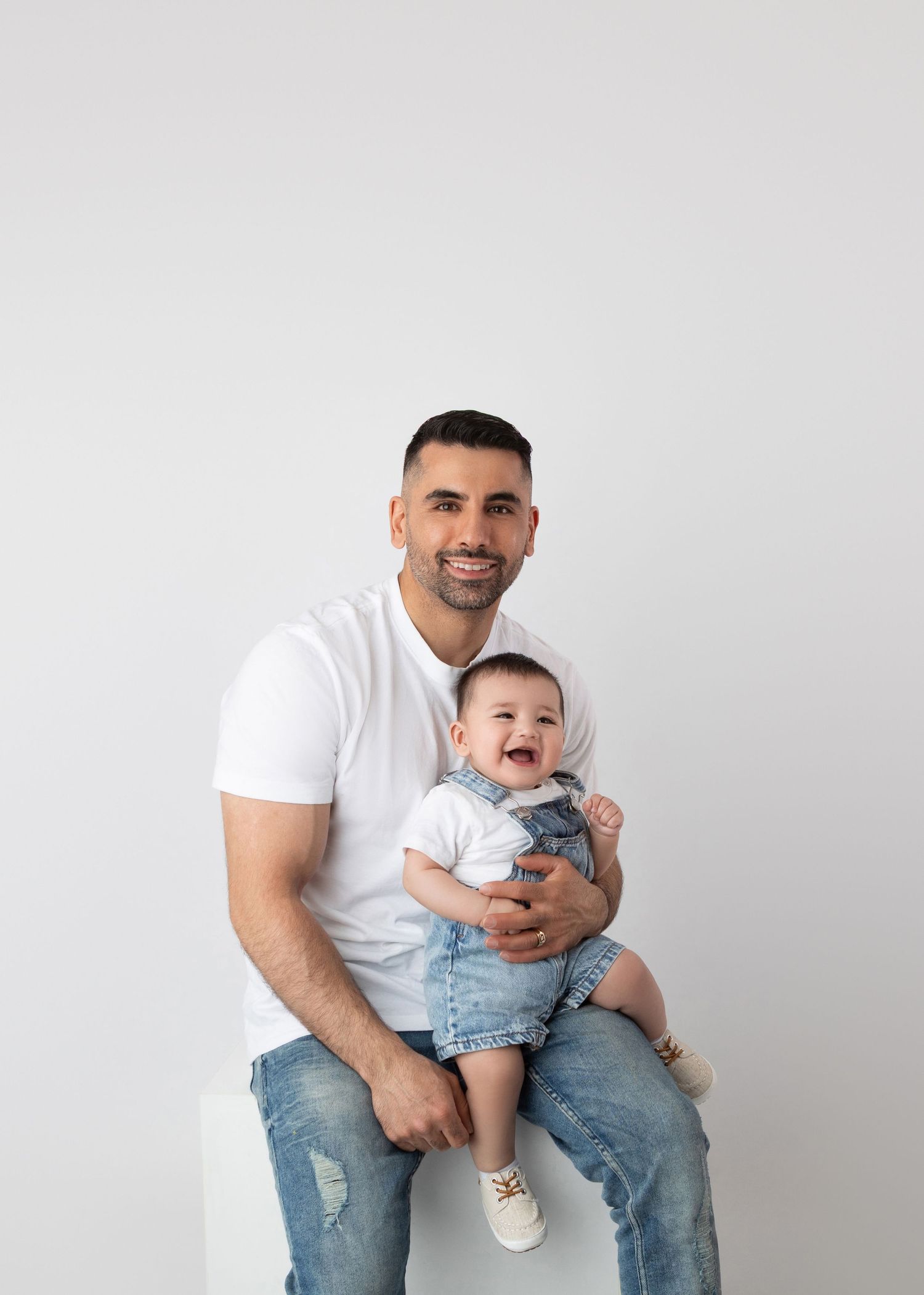 Man in white tee holding laughing baby in denim overalls, studio portrait.