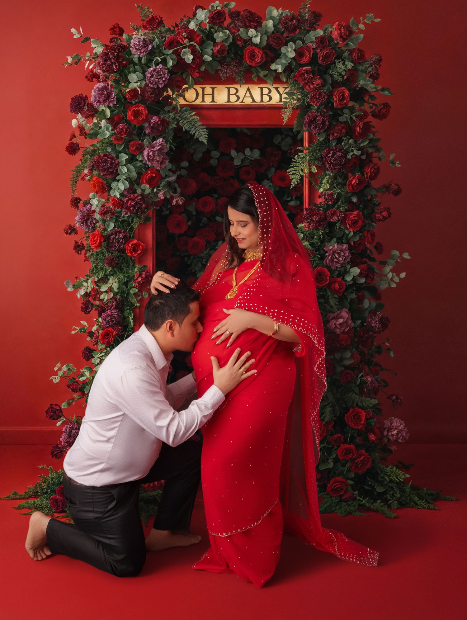 Couple in red maternity photoshoot, woman in red saree, man kneeling kissing belly, floral arch with OH BABY sign.