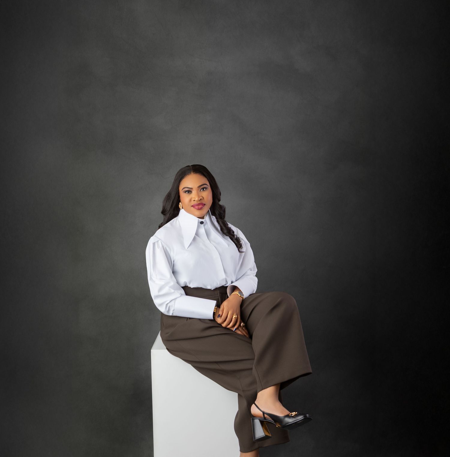 A confident woman in a white blouse and brown trousers sits on a white cube against a dark gray backdrop.