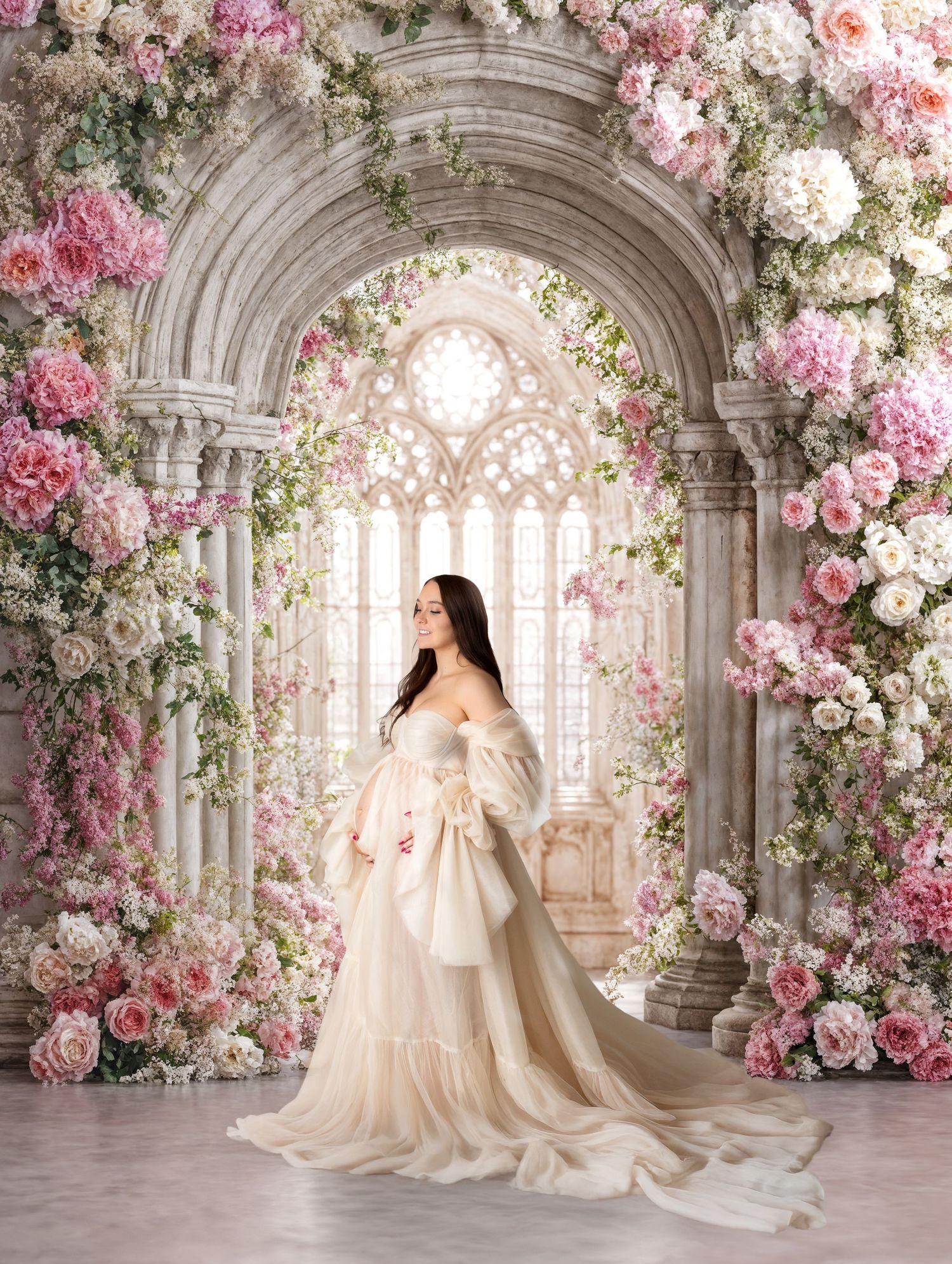 Pregnant woman in elegant cream tulle gown poses under floral arch with pink roses in dreamy studio setting.