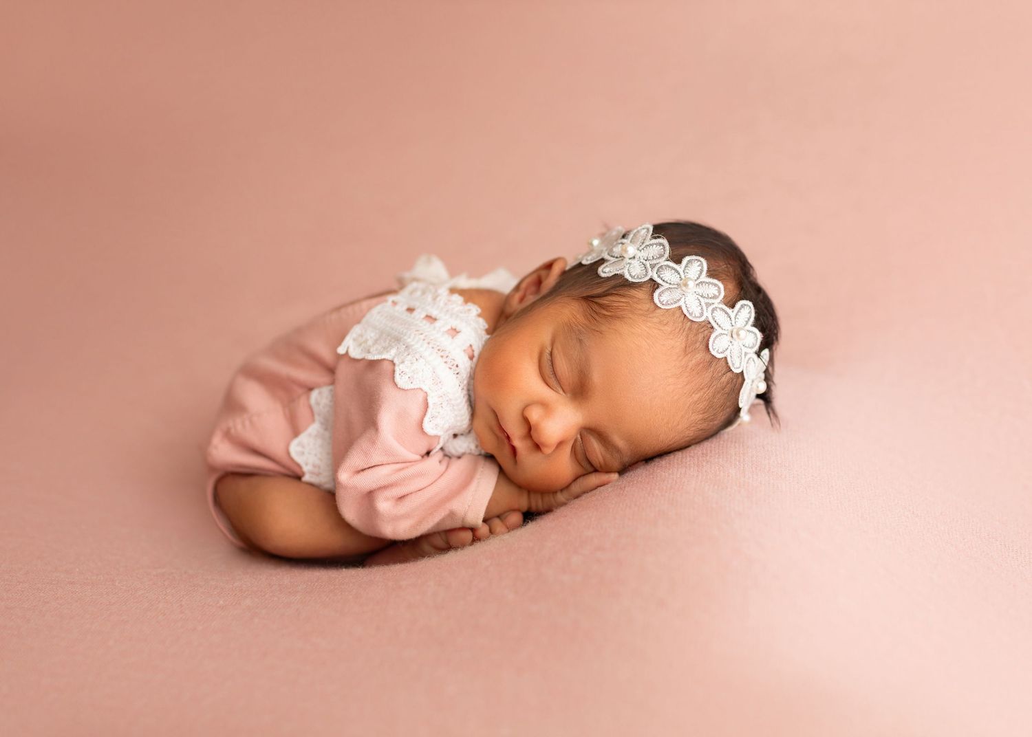 Sleeping newborn baby girl in pink romper with white lace headband on pink background.
