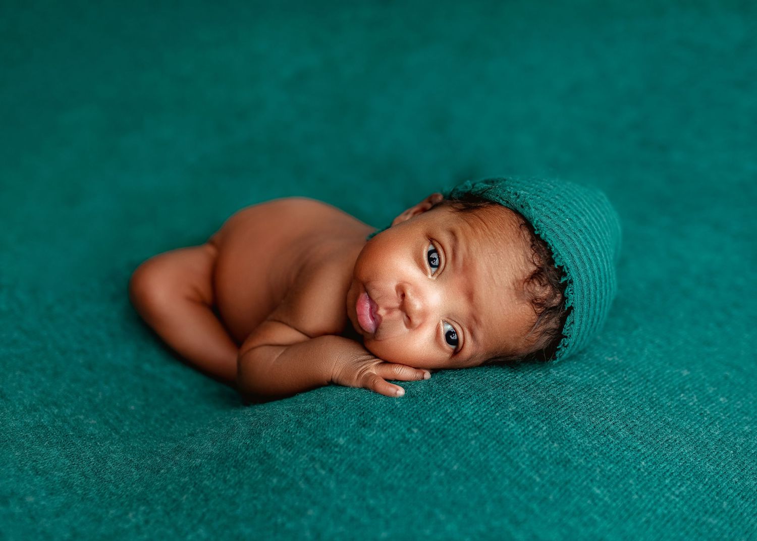 Awake newborn in a teal knit hat lying on a teal backdrop with a playful expression.