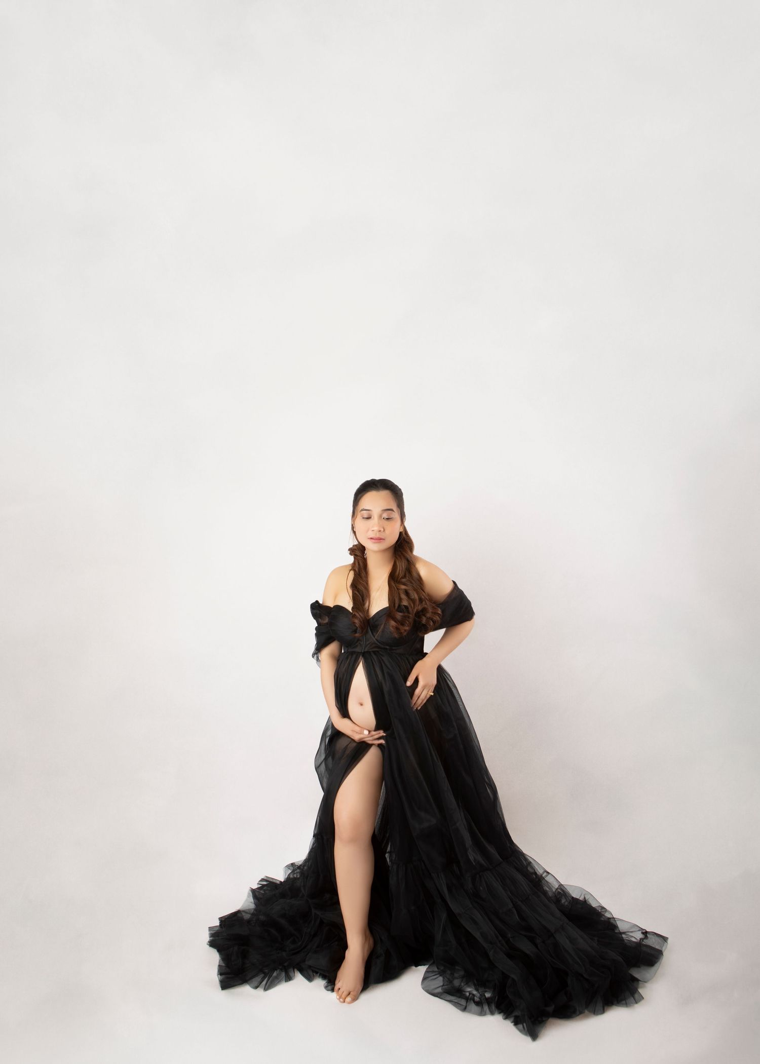 A pregnant woman in an elegant black ruffled tulle off-shoulder gown poses on a white background.
