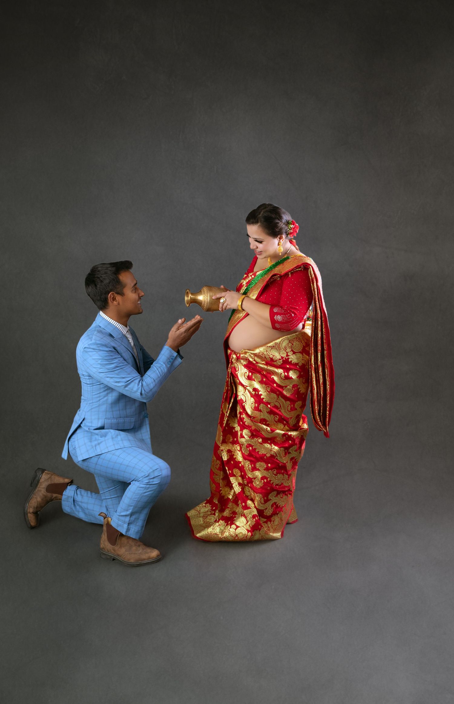 Man kneeling before pregnant woman in traditional red and gold saree during maternity shoot.