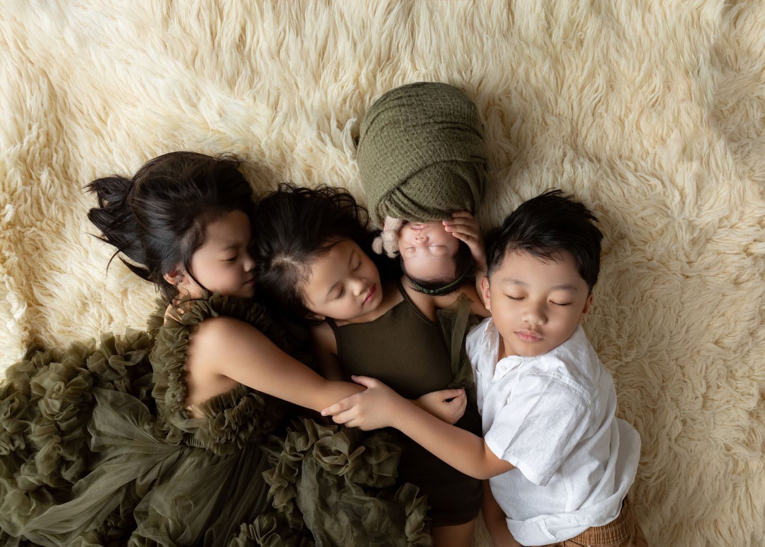 Three children cuddle together on a fluffy cream rug, with a swaddled newborn baby wrapped in green fabric.