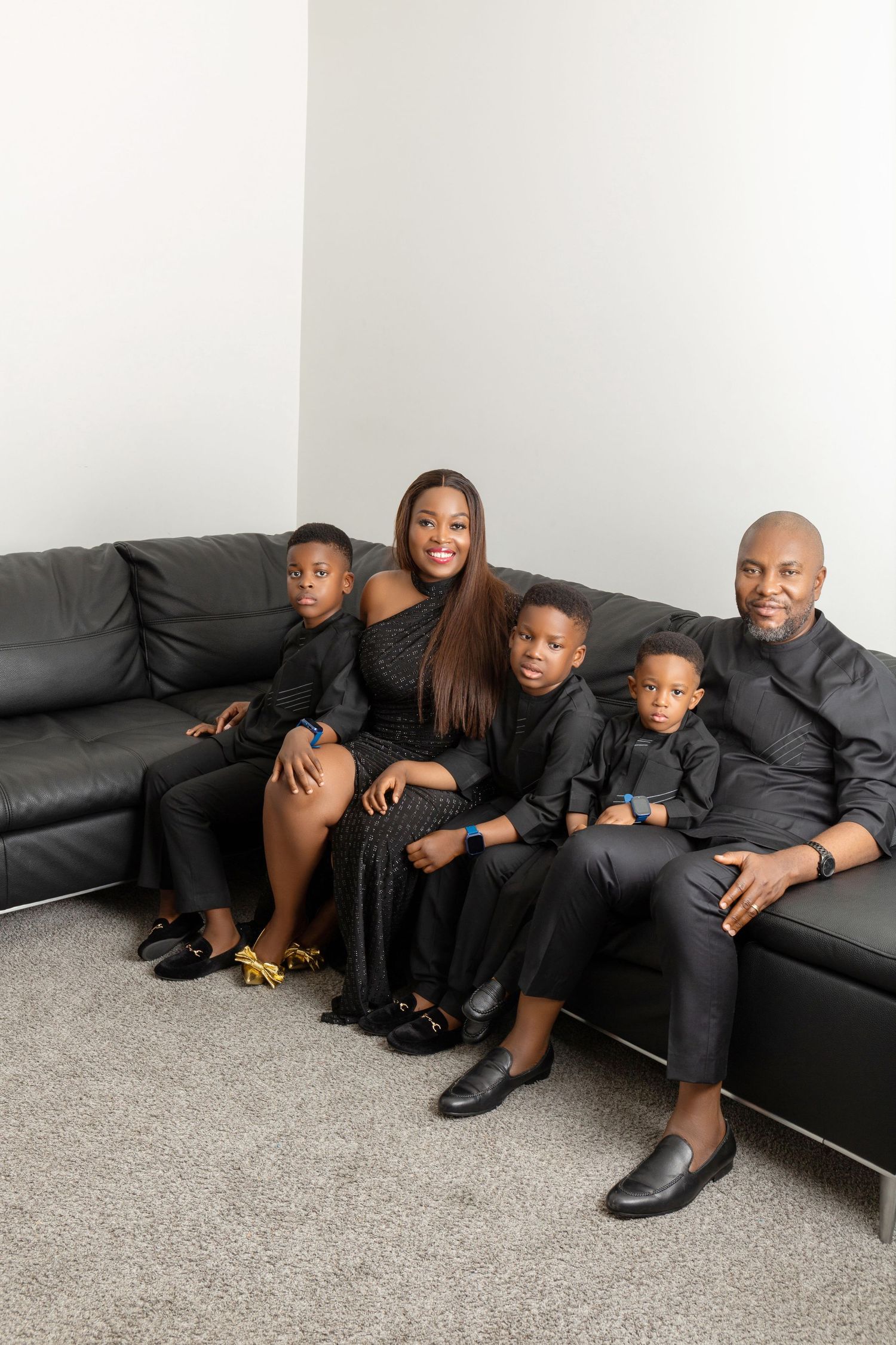 A Black family of five poses together on a black leather sofa, all dressed in matching black outfits.