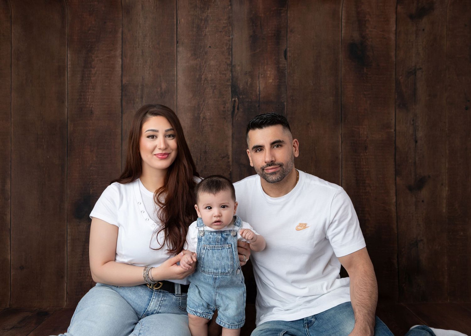 A young family poses for a portrait, with two parents in white tees and jeans holding their baby in denim overalls.