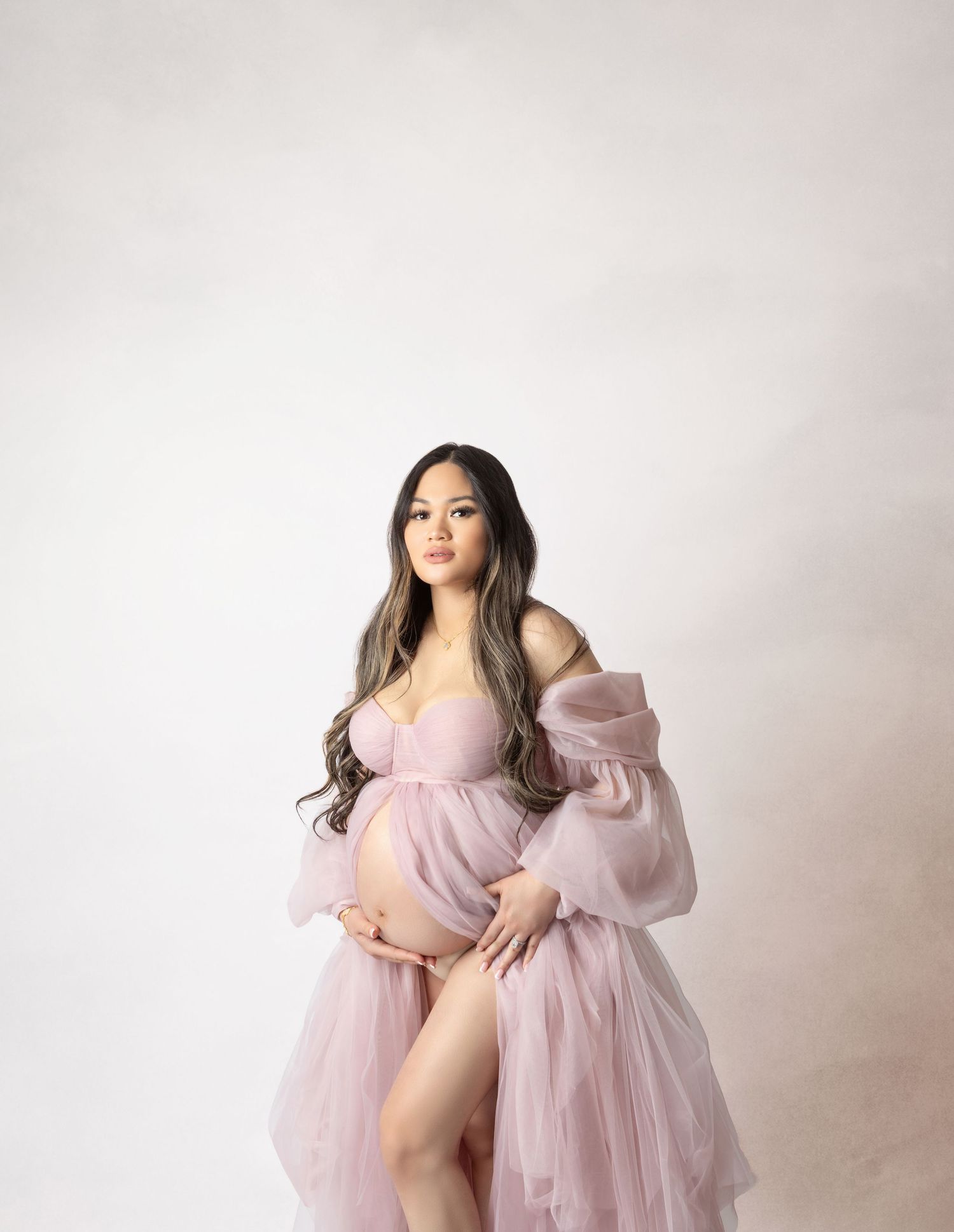 Pregnant woman in pink tulle maternity gown posing gracefully in a studio setting.