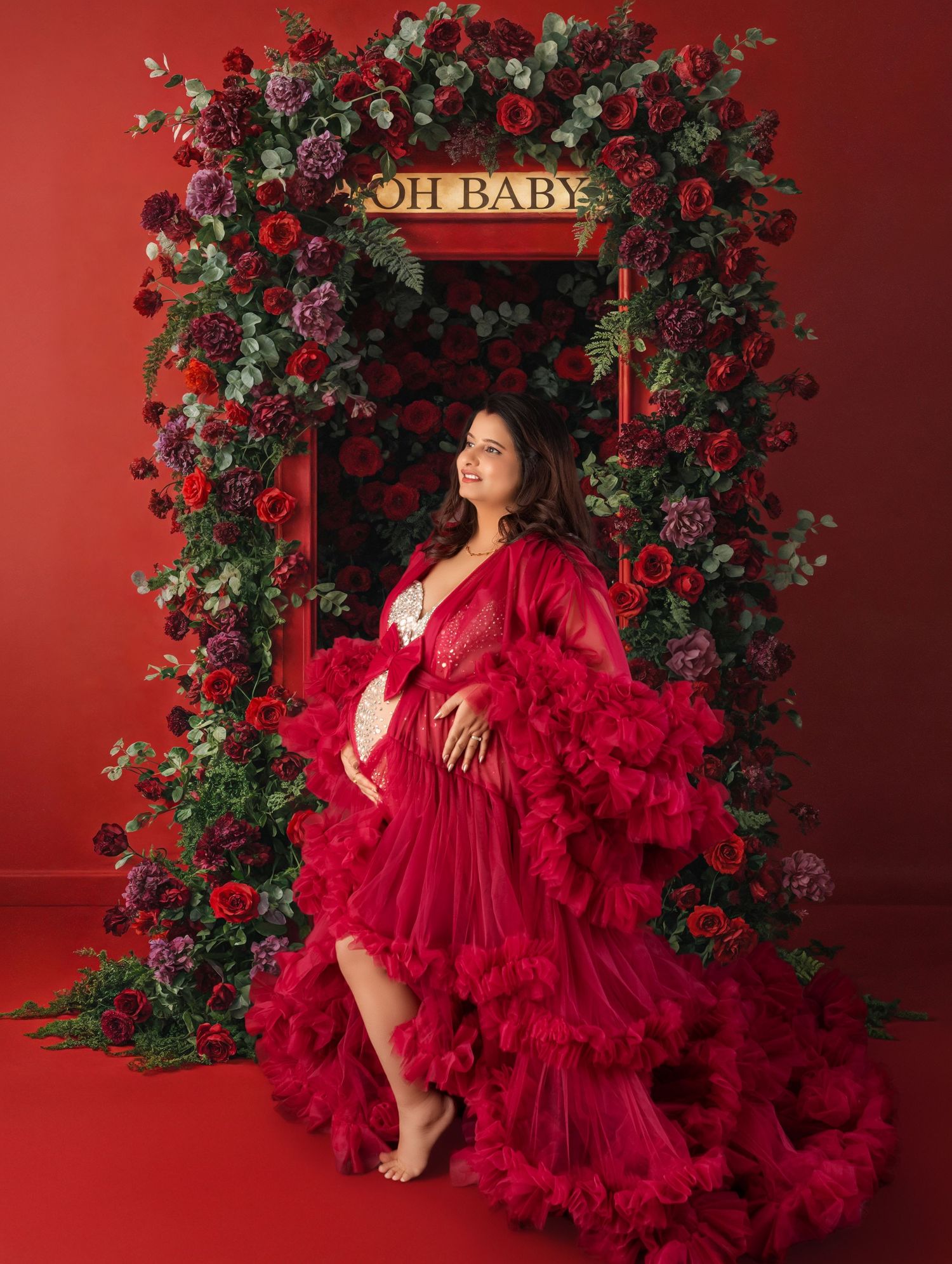 Pregnant woman in red tulle robe poses before floral arch with OH BABY sign for maternity photo.
