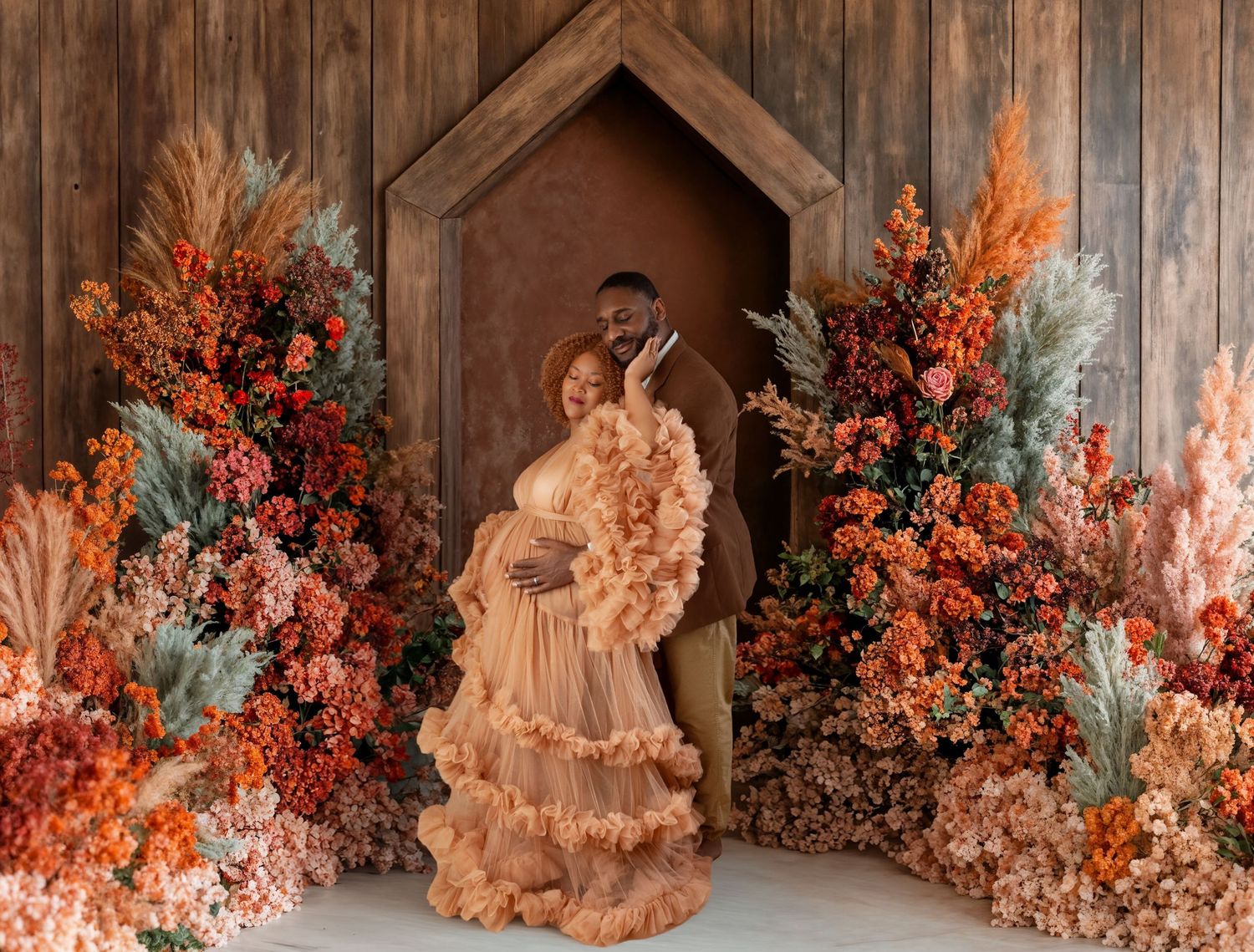Couple poses in maternity photo shoot surrounded by lush autumn floral arrangements in warm orange and red tones.
