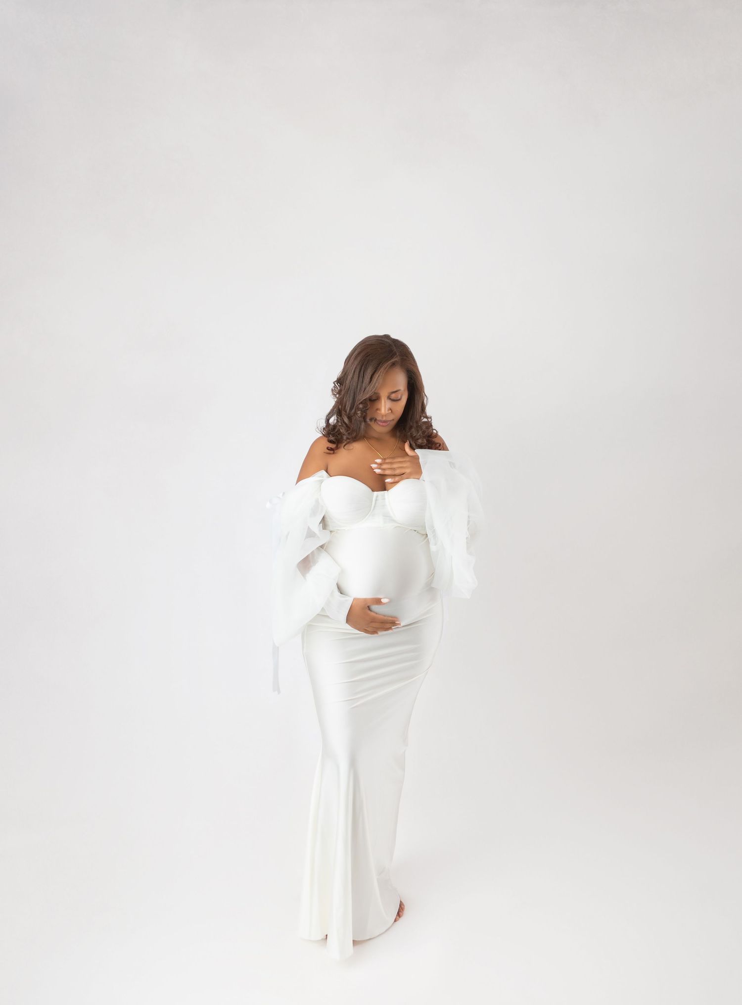 Pregnant woman in white off-shoulder fitted gown cradles baby bump in bright minimalist studio.