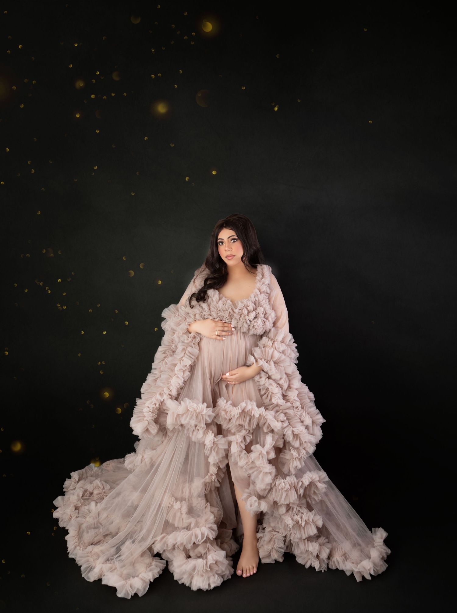Pregnant woman in blush pink ruffled tulle gown poses against dark backdrop with golden bokeh lights.