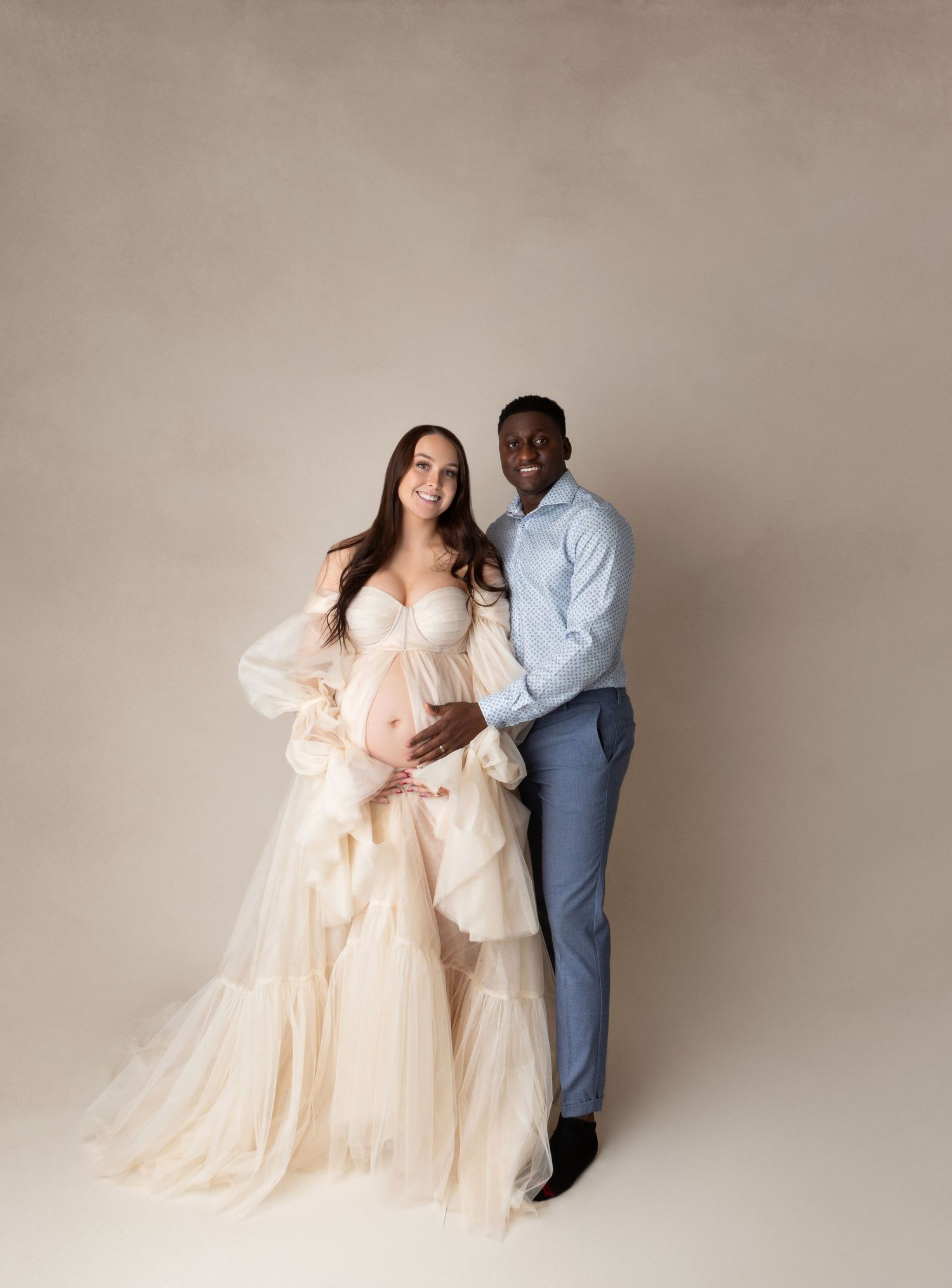 Smiling pregnant couple posing in studio, woman in cream ruffled tulle maternity gown, man in light blue suit beside her.