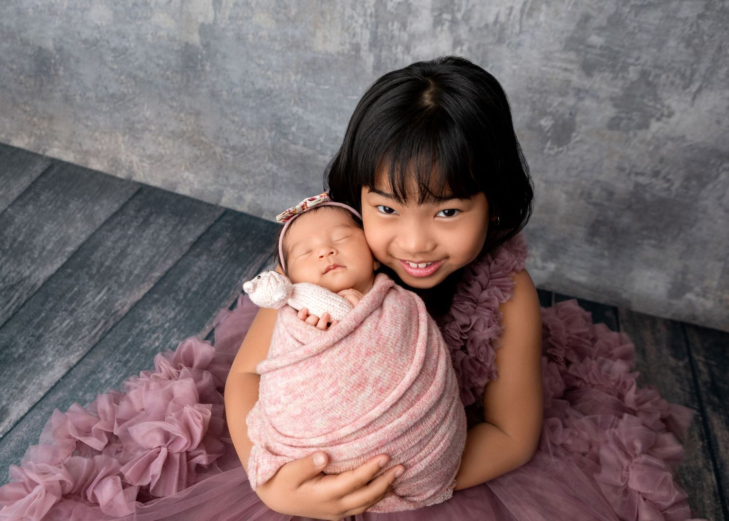Smiling young girl proudly holds swaddled newborn baby sister in purple ruffle dress portrait.