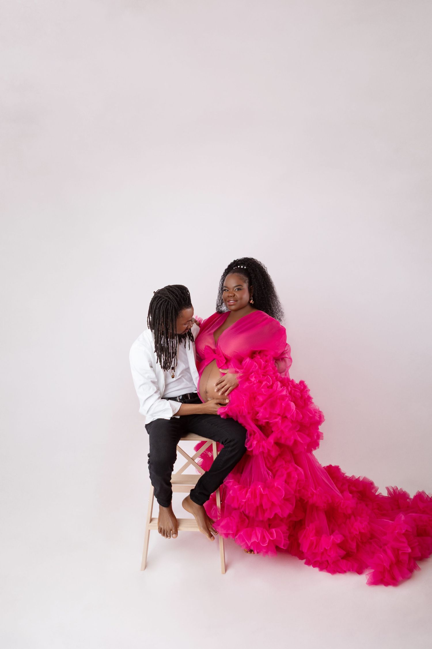 Couple sharing a tender moment during maternity photo shoot, woman in pink ruffled gown on white background.