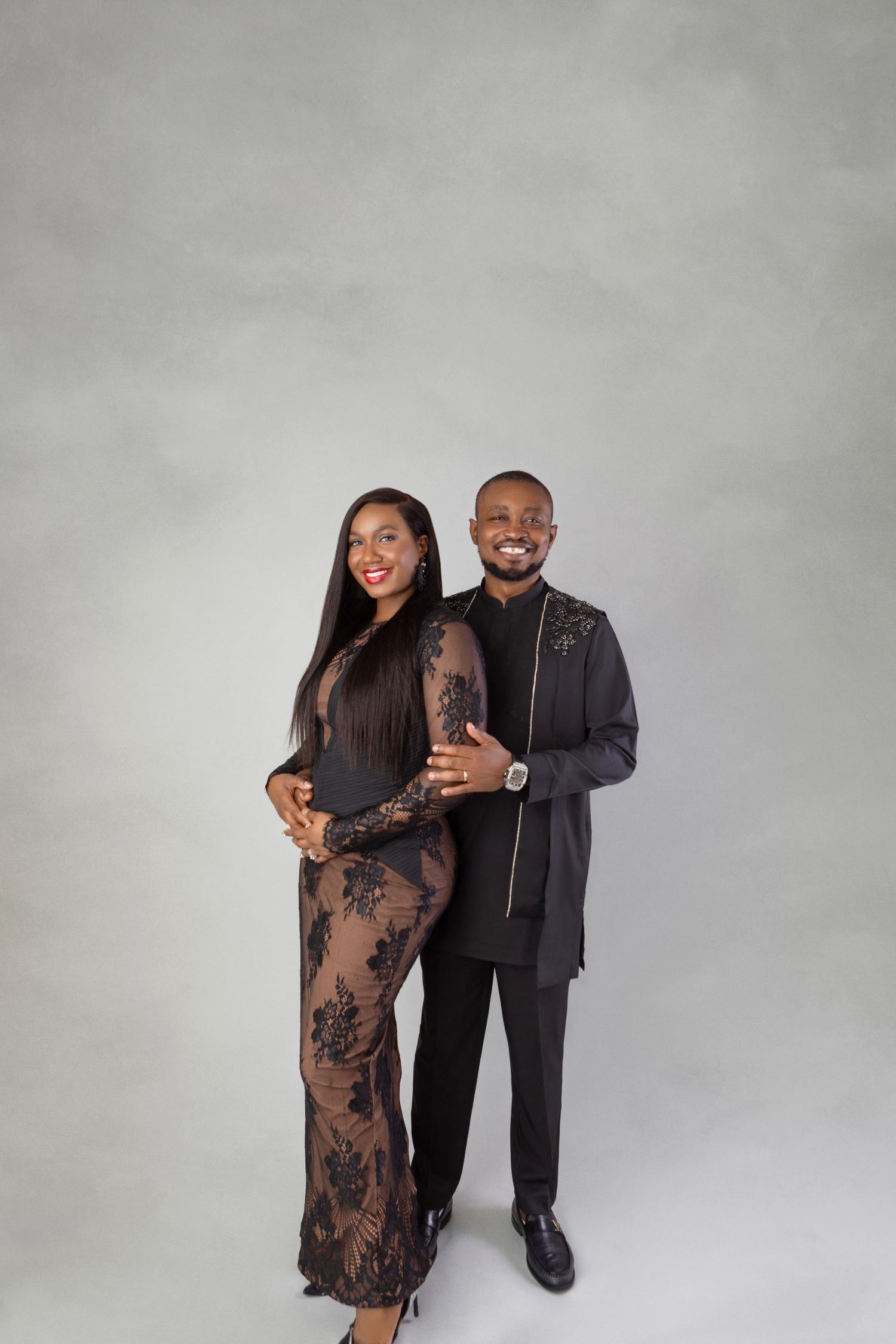 Couple in coordinating black outfits embrace and smile against gray studio background.