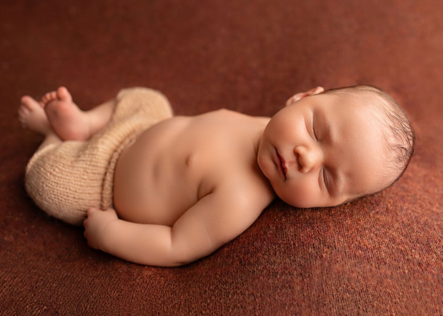 Peaceful newborn baby sleeping curled up on a warm brown blanket, wearing knit tan shorts.