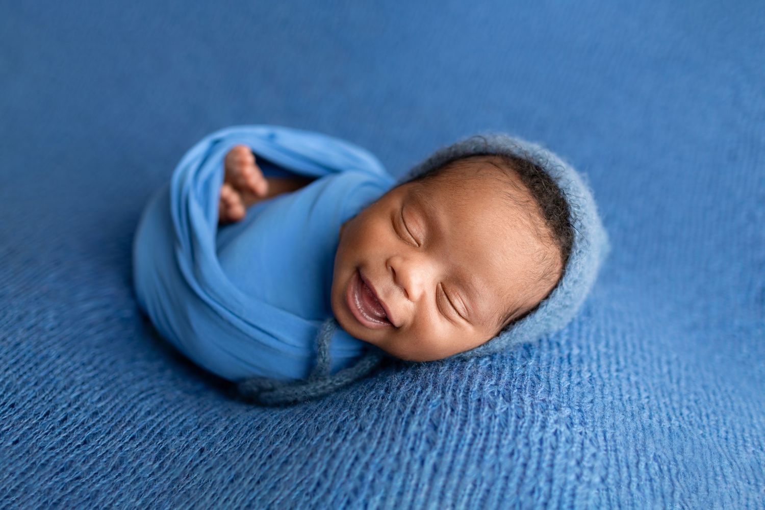Smiling newborn baby wrapped in blue swaddle with knit bonnet on blue background.