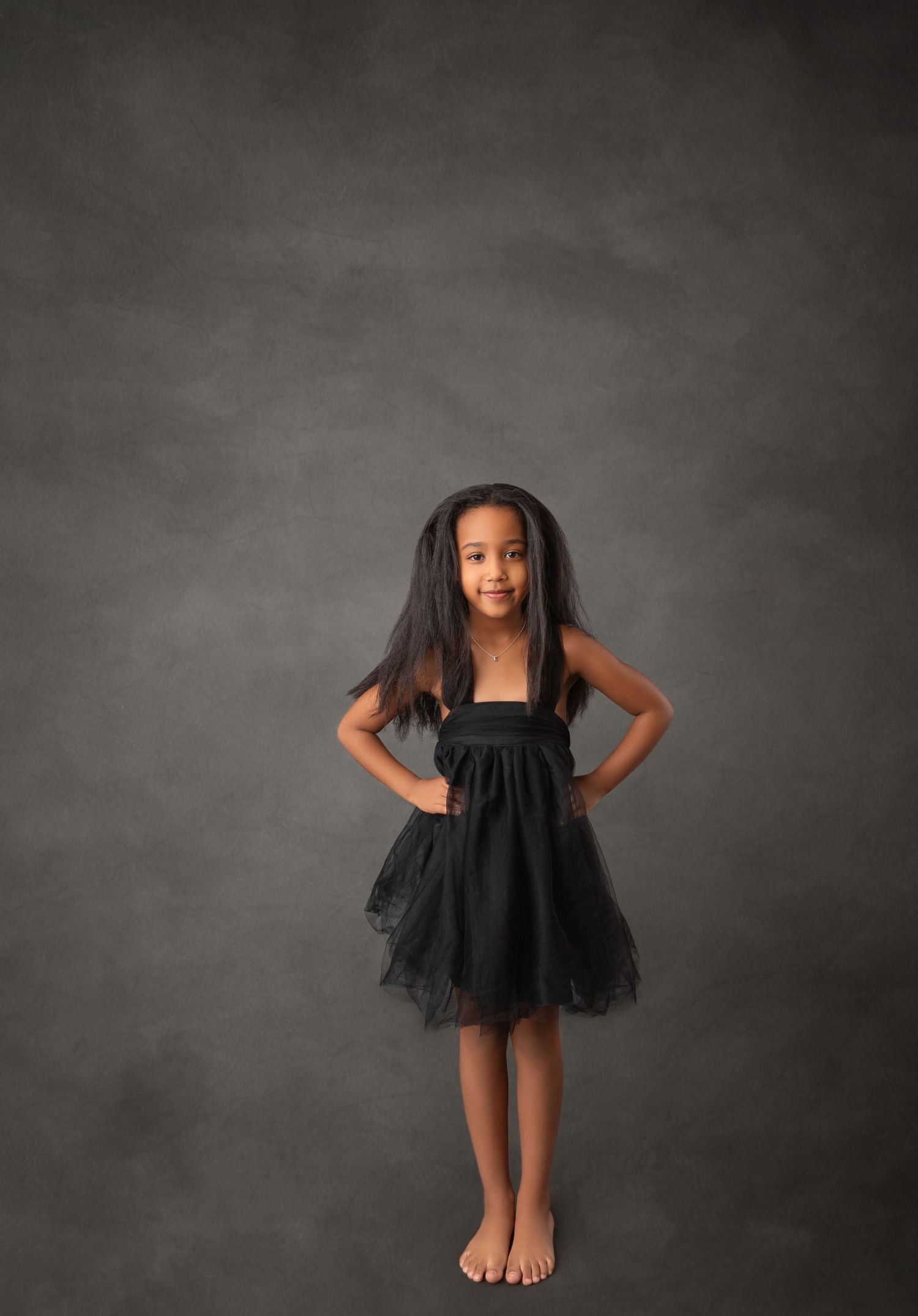Young girl in black tulle dress poses barefoot against gray backdrop, hands on hips, long dark hair flowing.