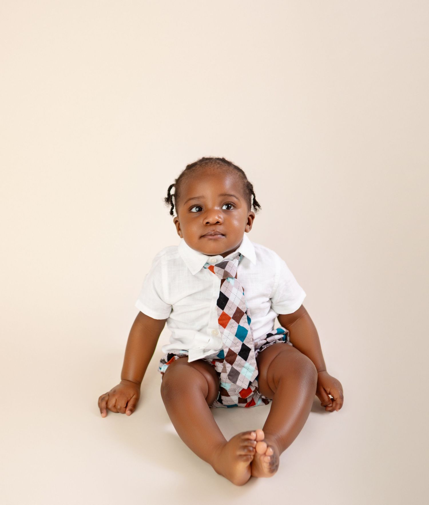 Adorable Black baby boy sitting in white shirt and colorful argyle tie, looking up curiously against beige background.