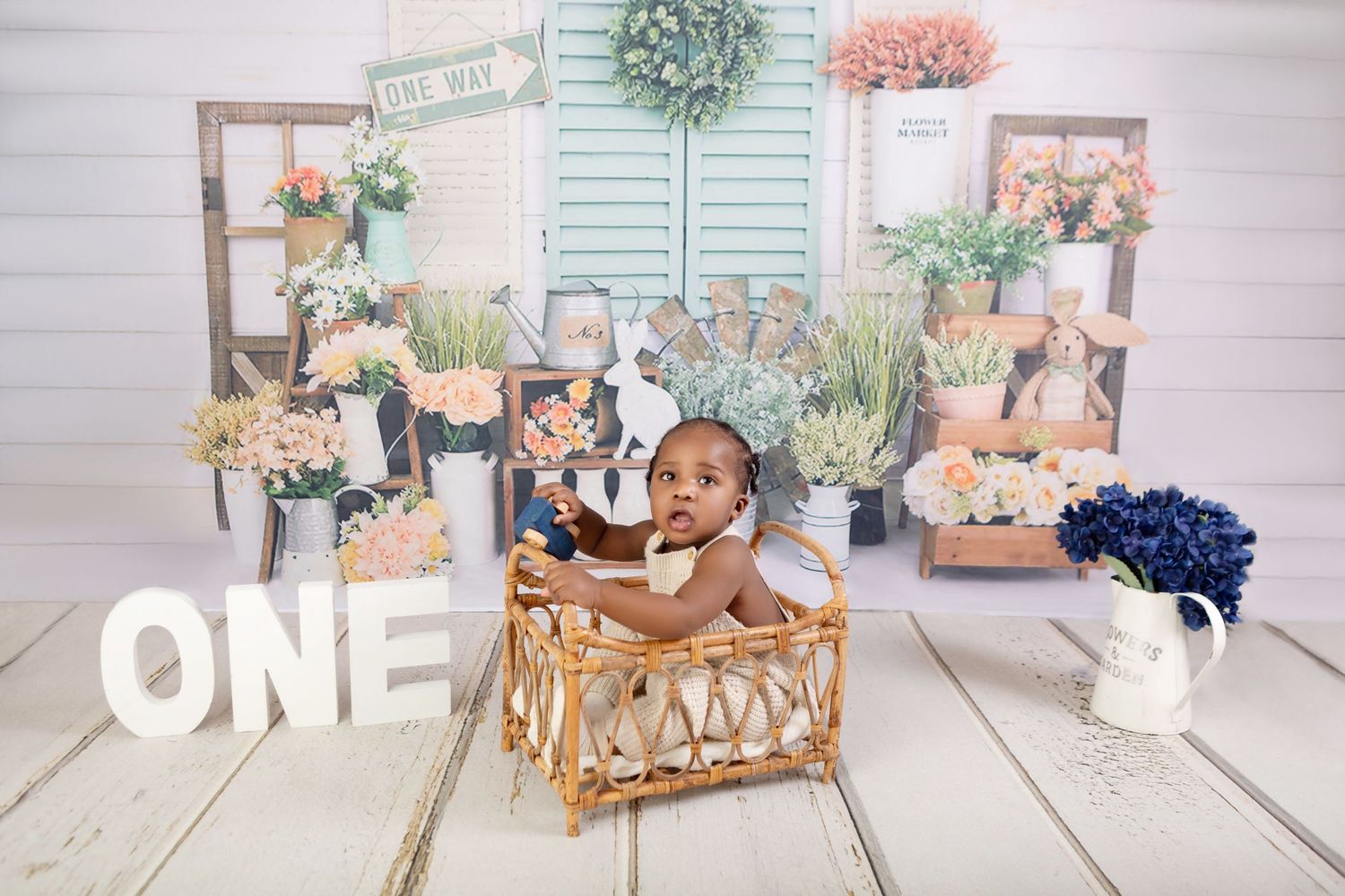 Baby's first birthday photo shoot with floral backdrop, wicker basket, and ONE sign on white wood floor.