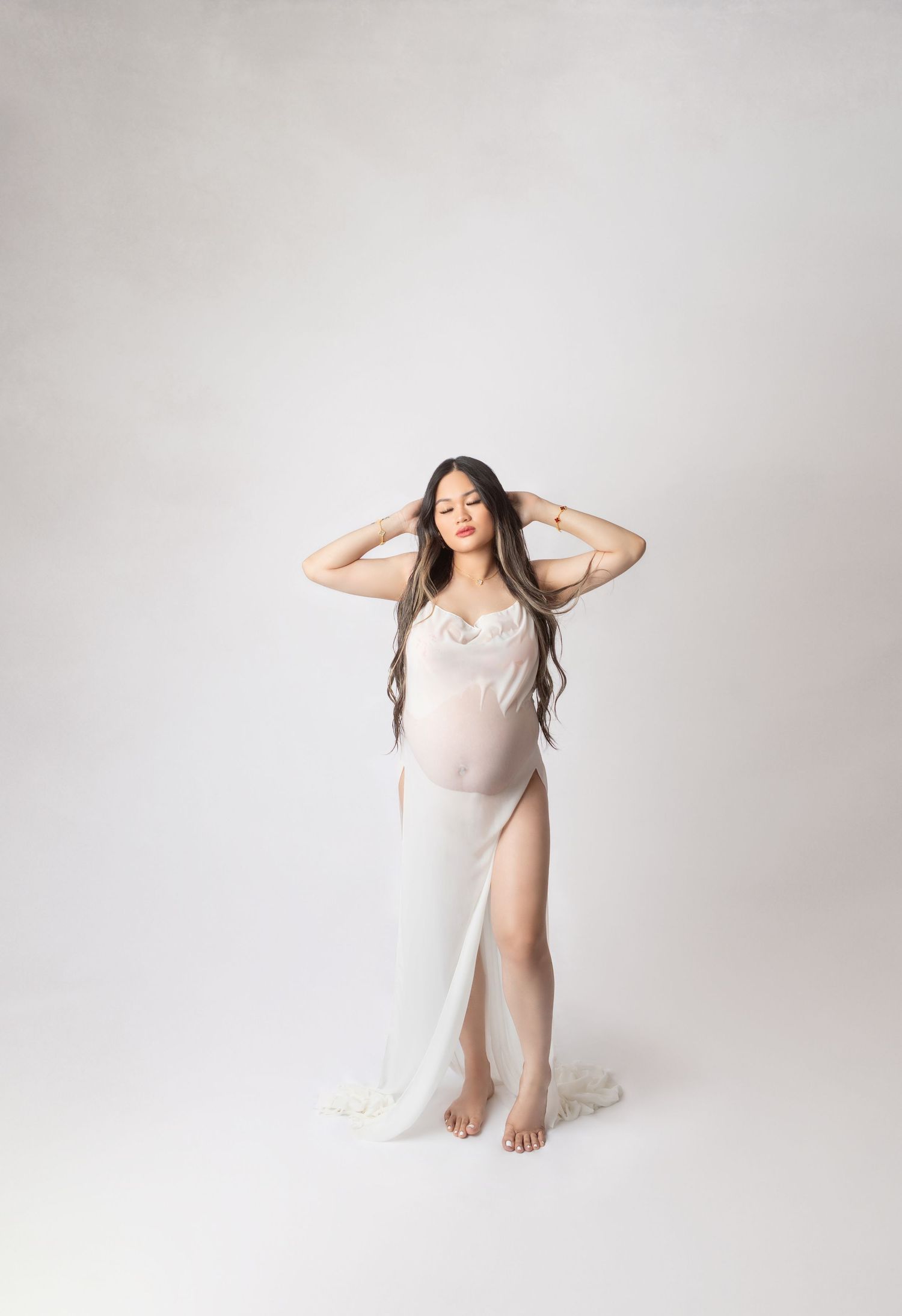 Pregnant woman in sheer white maternity gown poses gracefully in a bright studio setting.