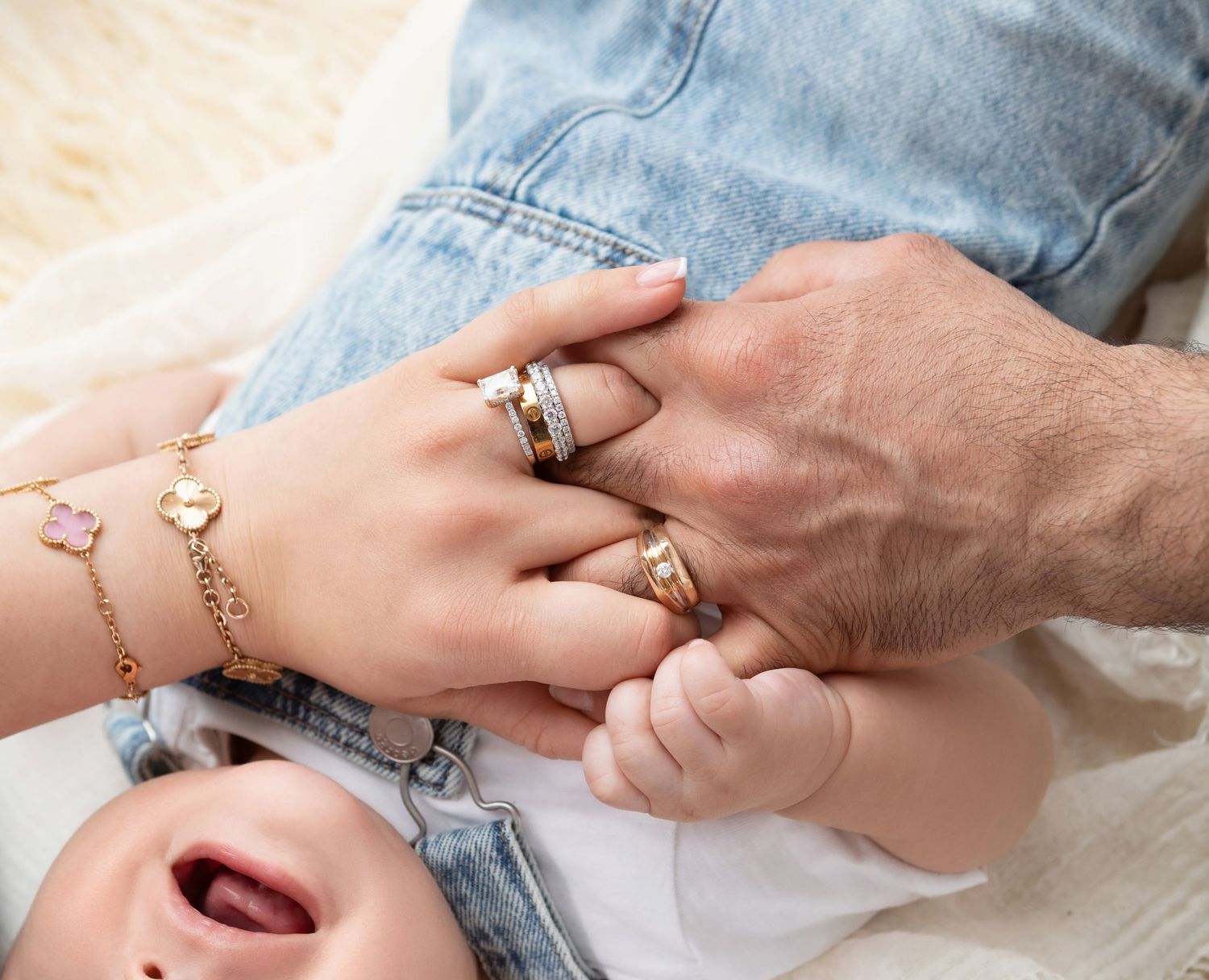 A baby holds parents' hands adorned with gold rings and bracelets, lying on a soft white blanket.
