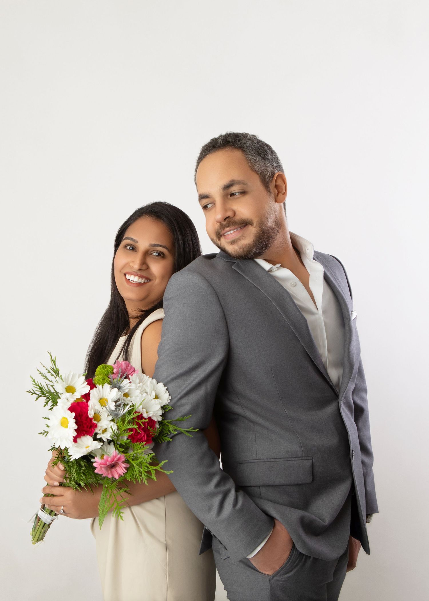 Smiling couple posing back-to-back, woman holding colorful flower bouquet, man in gray suit, white background.