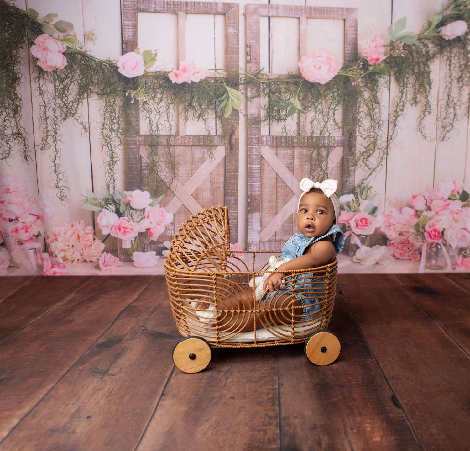 Baby girl in wicker pram with floral backdrop and barn doors for a charming photo shoot.