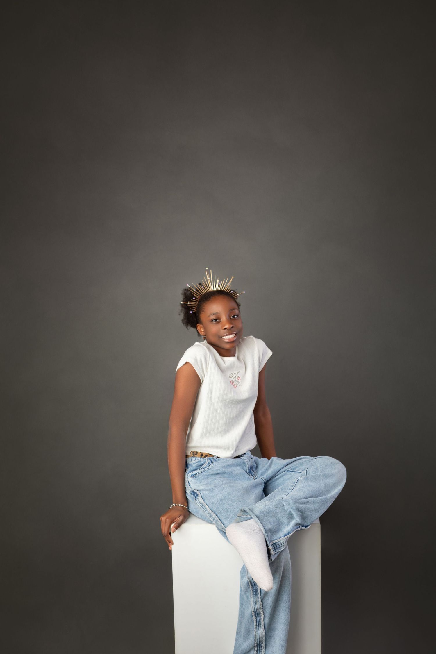 Smiling young Black girl in white top and jeans sitting on white cube against gray background.