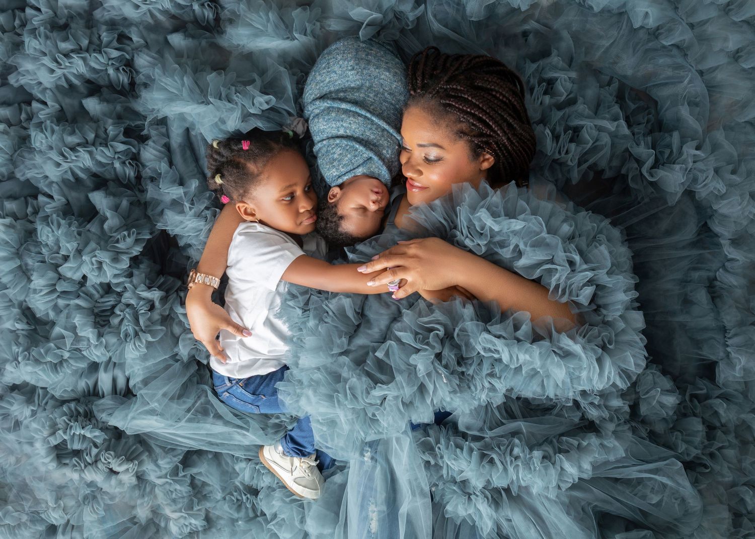 Mother lying on blue tulle fabric smiling at her two young children during a professional photo session.