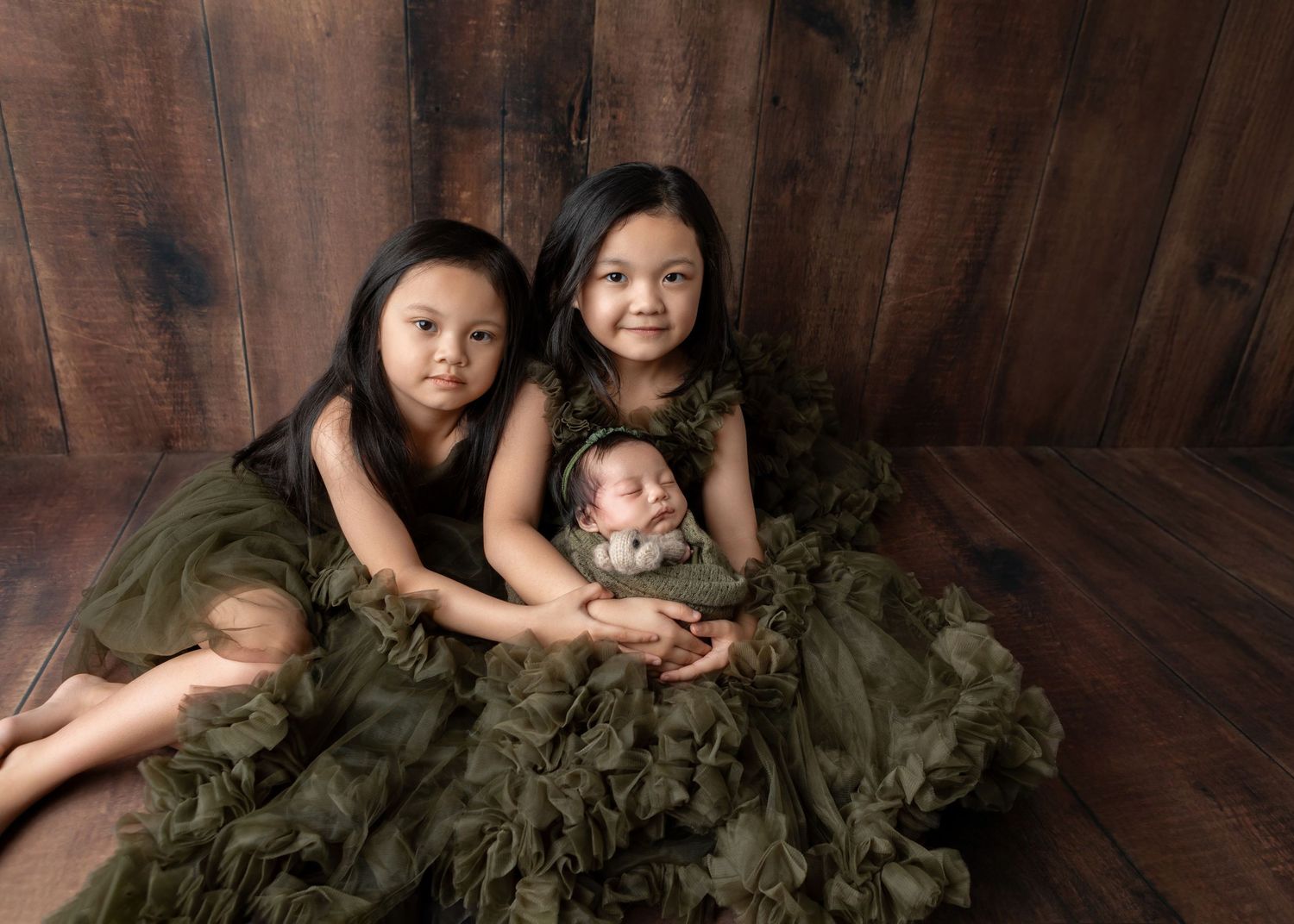 Two girls in olive green tulle dresses pose with a sleeping newborn baby wrapped in matching green fabric.