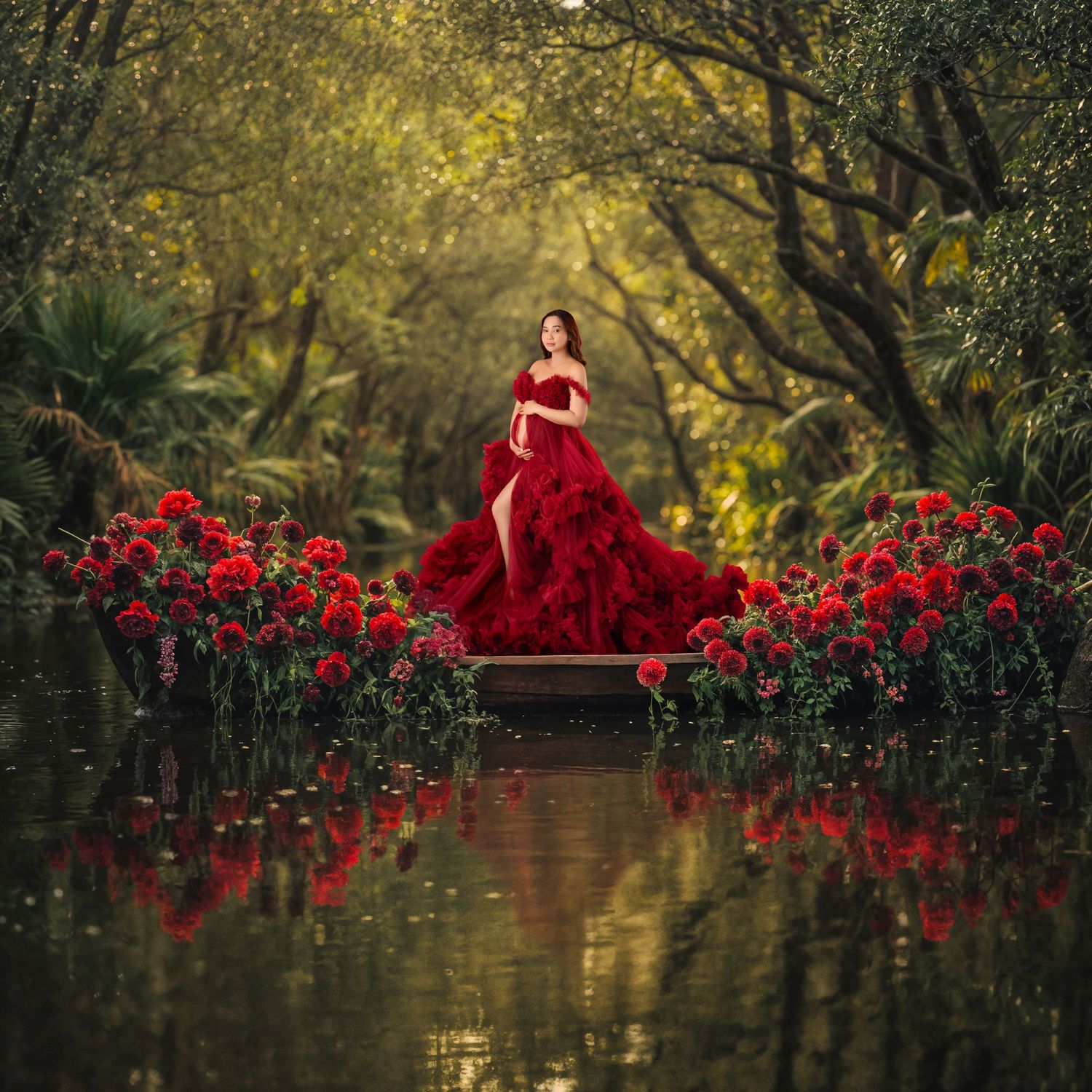 Woman in red gown stands on dock surrounded by red flowers on a tranquil forest waterway.