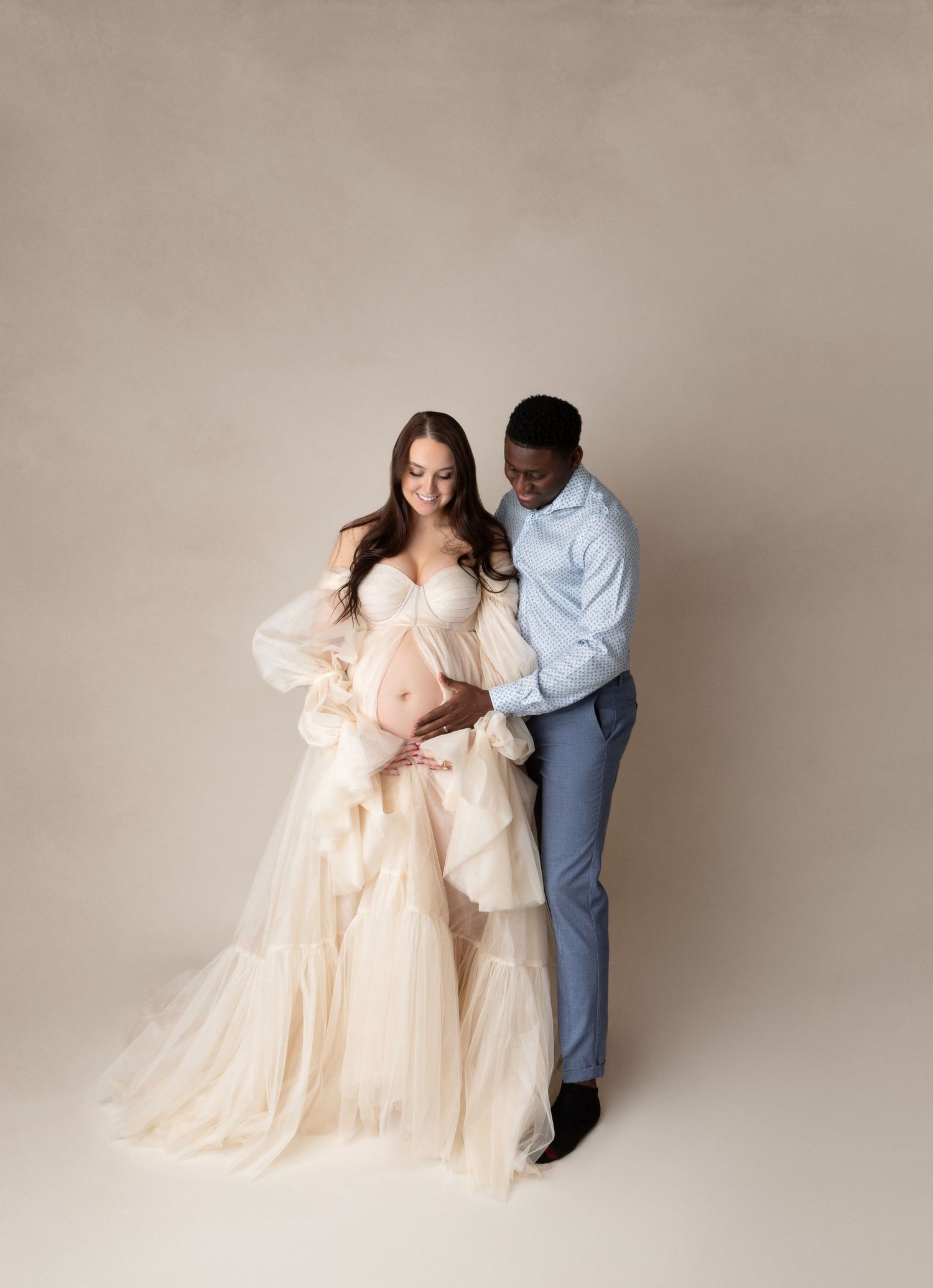 Expecting couple sharing tender moment in studio, woman in elegant cream tulle maternity gown, man in blue suit cradling her bump.