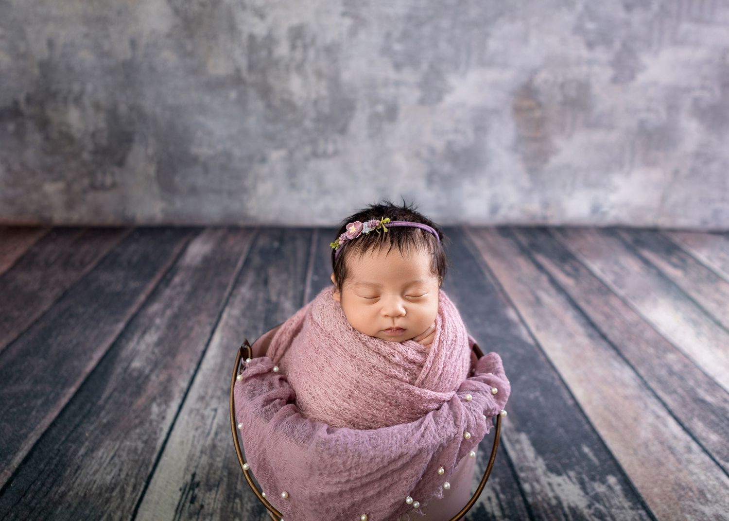 Sleeping newborn baby girl swaddled in pink wrap with floral headband in wooden bowl prop.