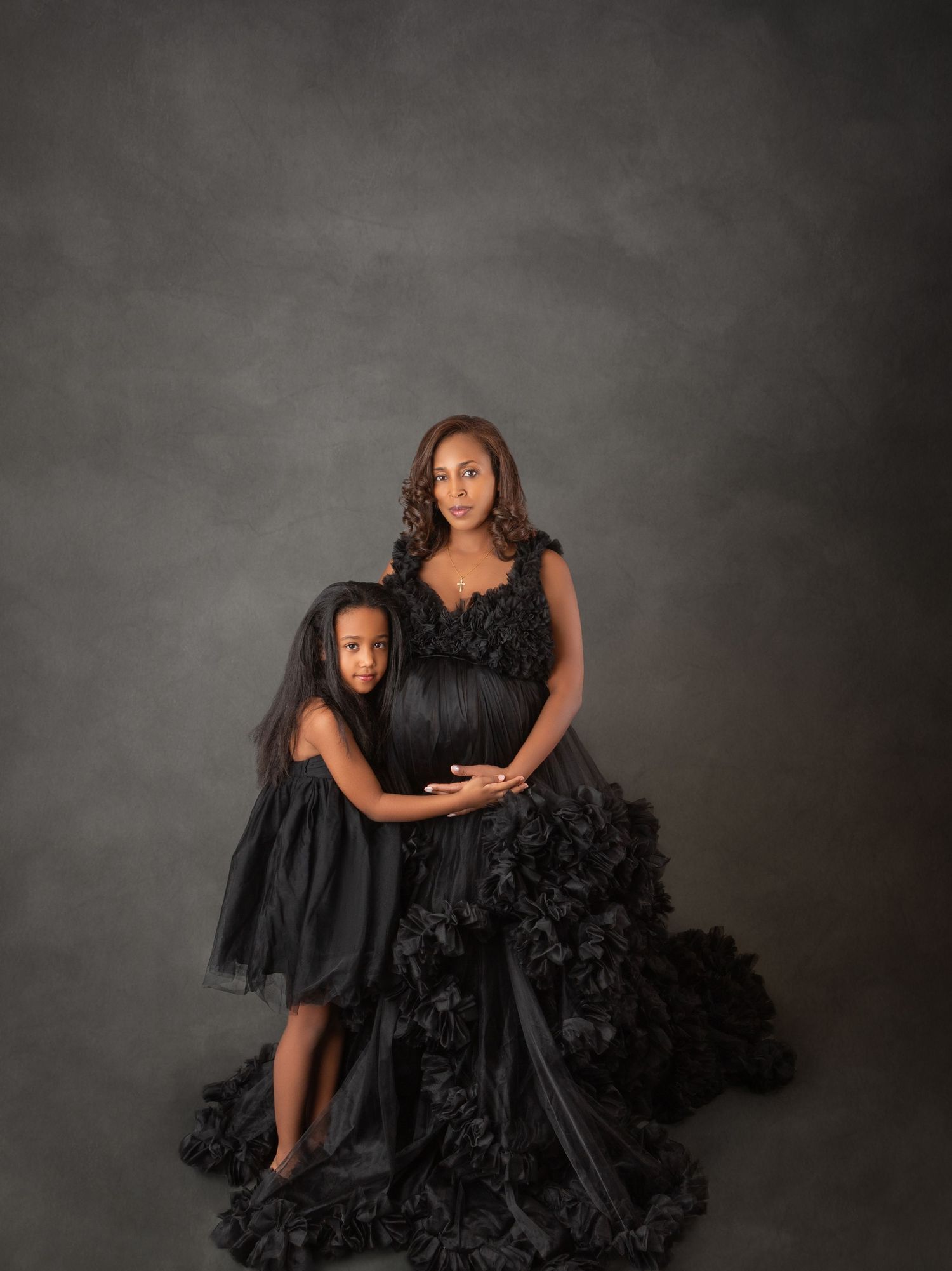 Pregnant mother and daughter wear matching black ruffled gowns in elegant studio maternity portrait.