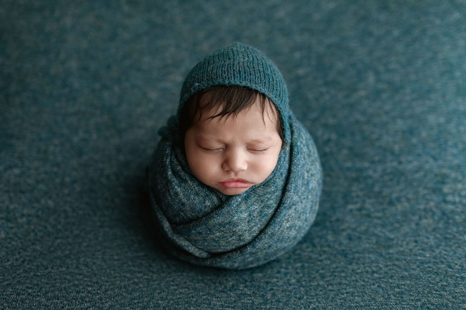 Sleeping newborn baby wrapped in teal knit swaddle and matching hat posed on a teal textured blanket.