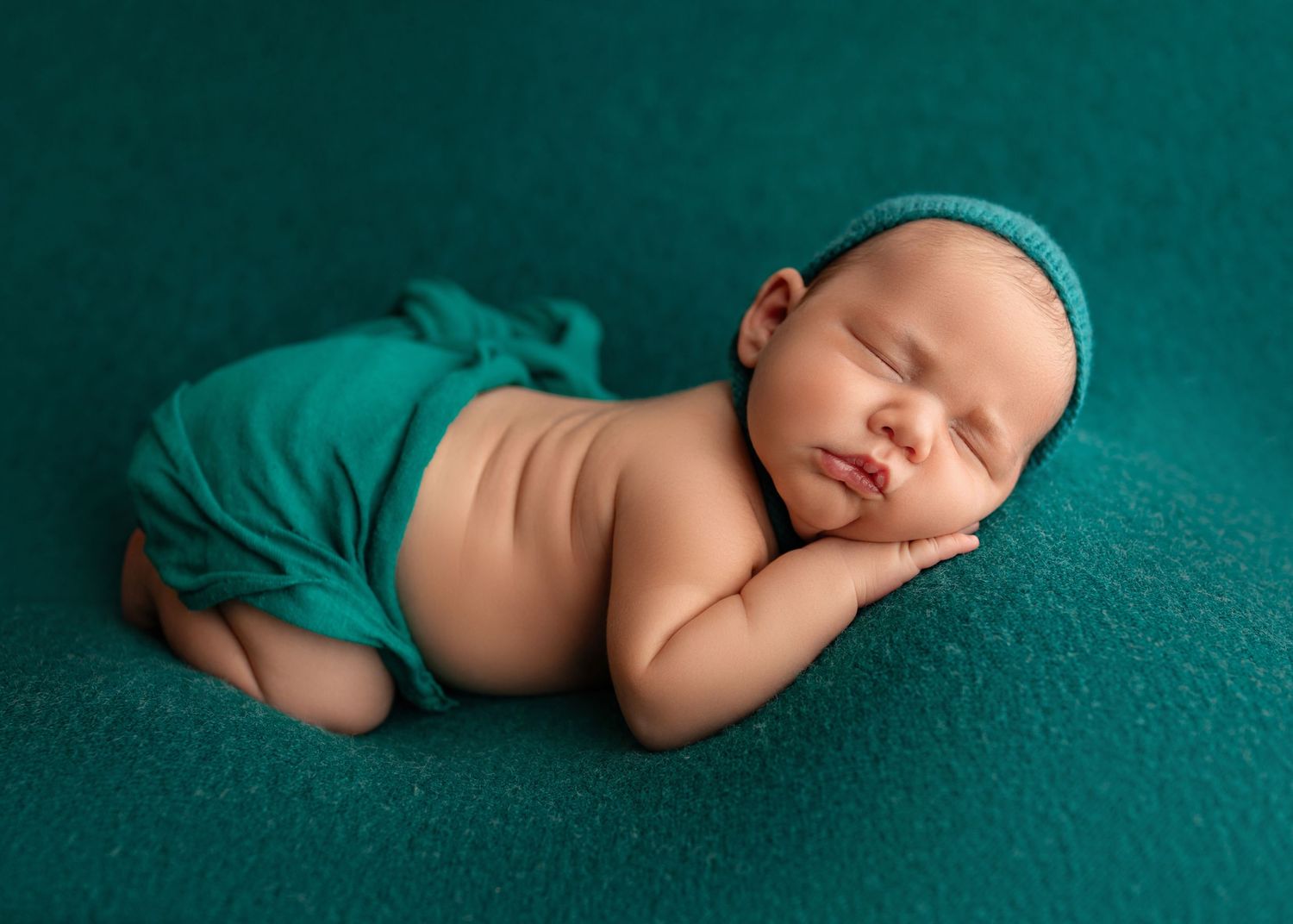 A peaceful newborn baby sleeping curled up on a teal blanket, wearing a matching knit hat and wrap.