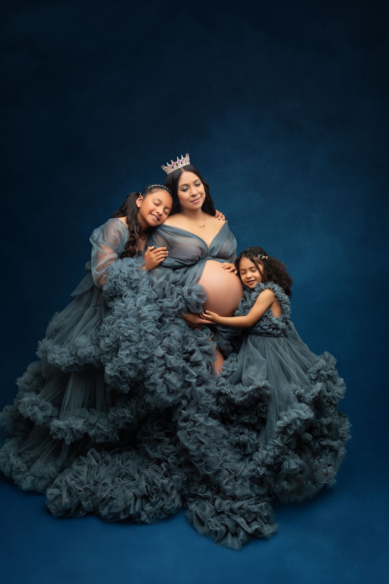 Pregnant woman wearing crown poses with two daughters in matching dusty blue ruffle tulle gowns.