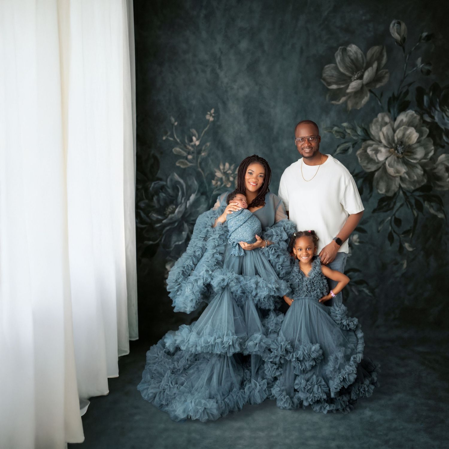 Family portrait with mother, father, and two children in elegant blue tulle gowns against floral backdrop.